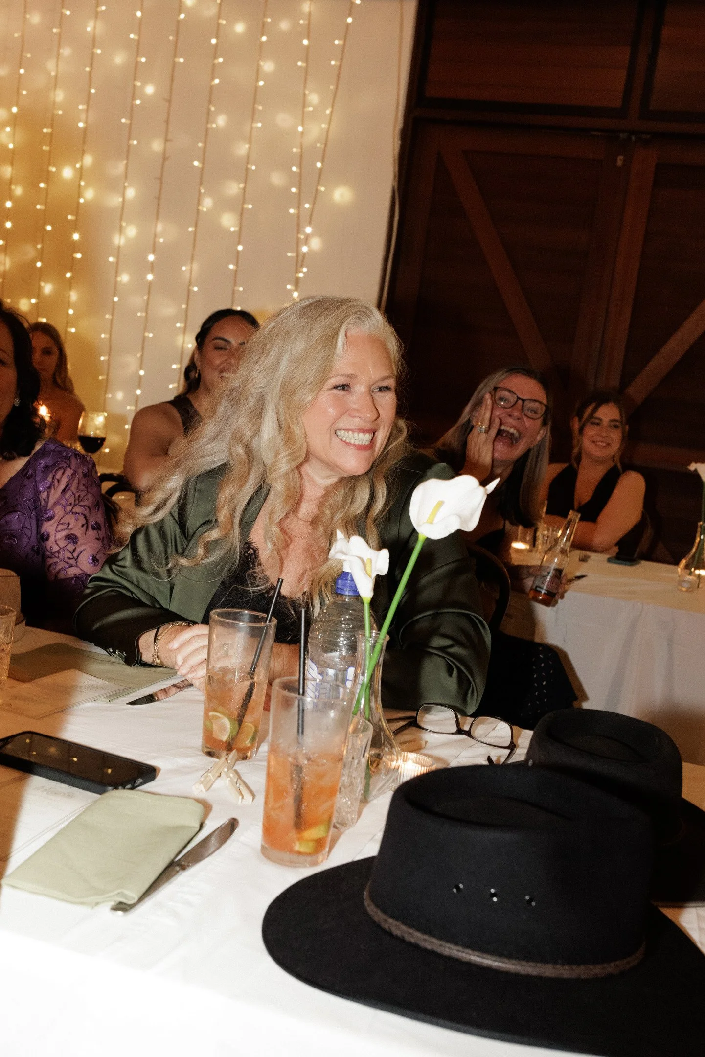 People gathered at a dining table in a warmly lit room, with string lights hanging on the wall in the background. A woman with blonde hair in the foreground is smiling, and someone nearby is wearing glasses and laughing. The table has drinks, glasses