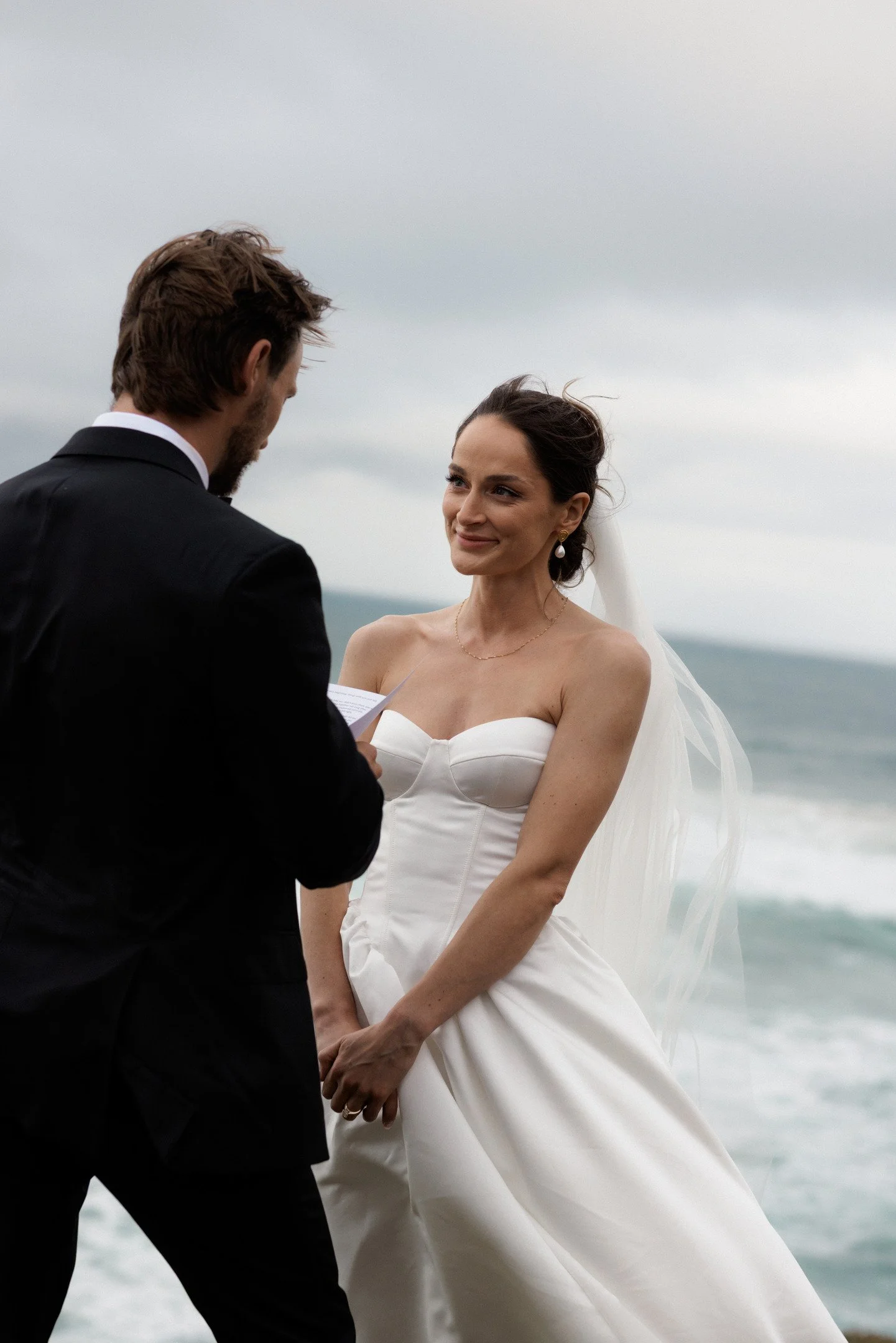 A bride and groom stand facing each other outdoors during their wedding ceremony, with a cloudy sky and body of water in the background.