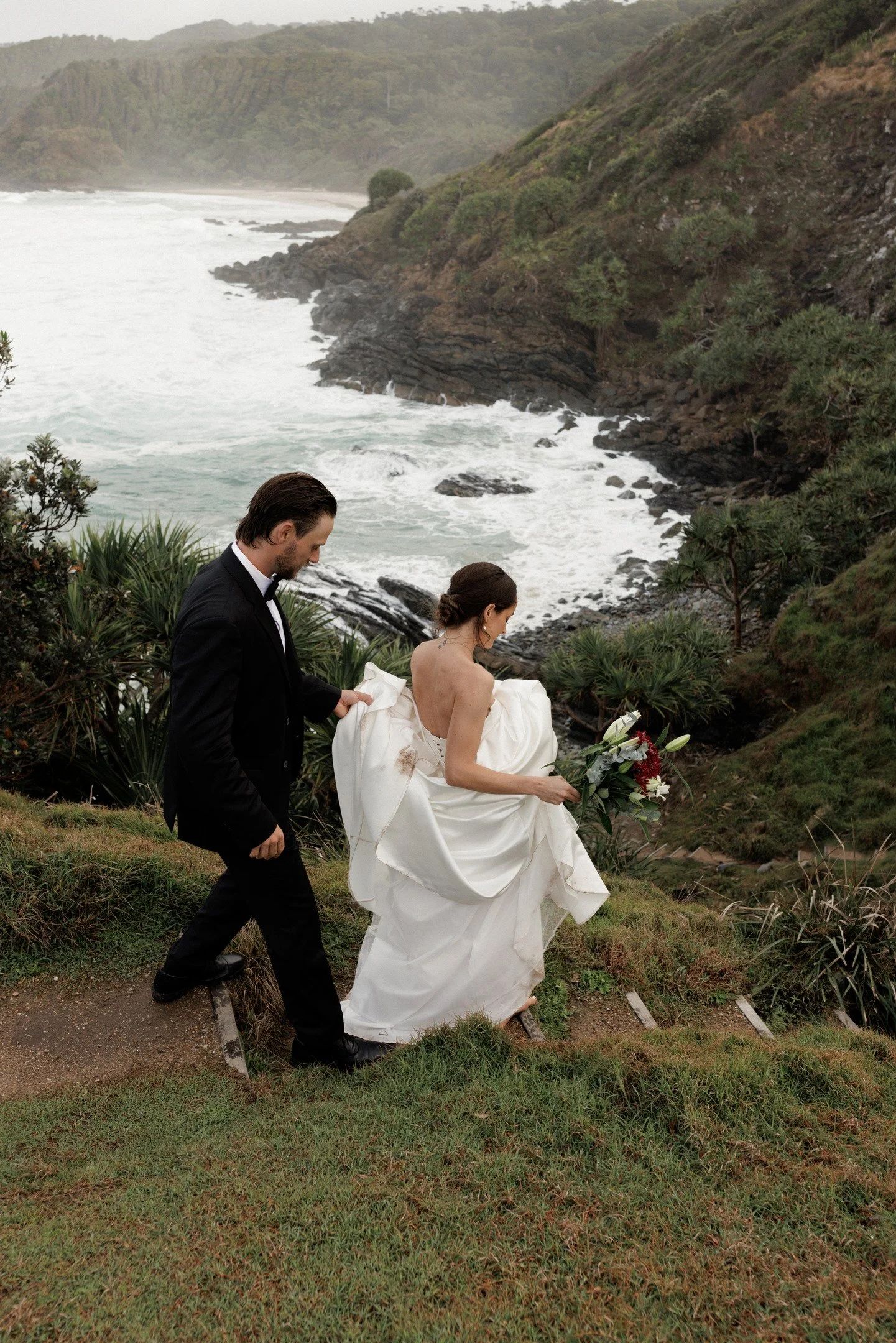 A man and woman dressed in formal attire, possibly for a wedding, walking along a coastal trail with cliffs, ocean waves, and lush greenery in the background.