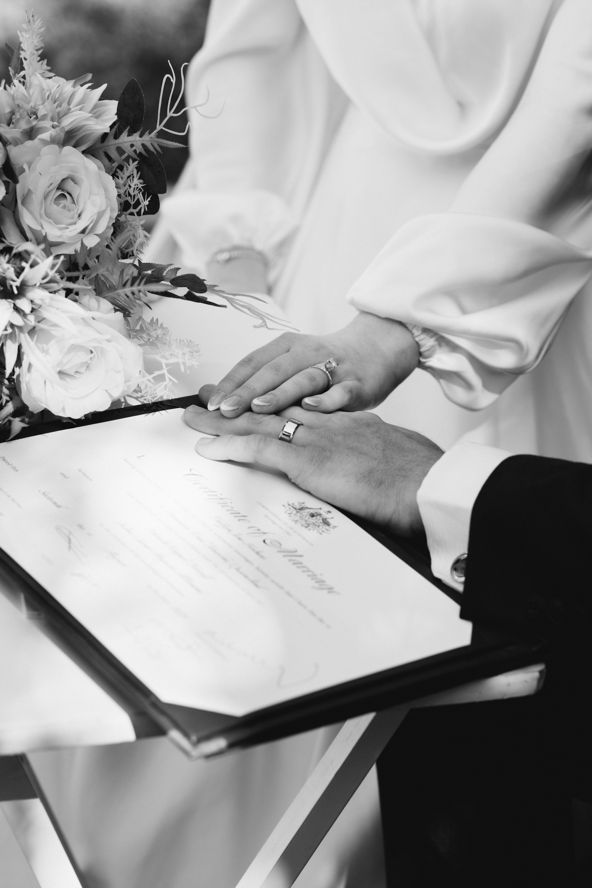 Close-up of a couple's hands, wedding rings visible, resting on a marriage certificate during a wedding ceremony, with a bouquet of flowers nearby, in black and white.