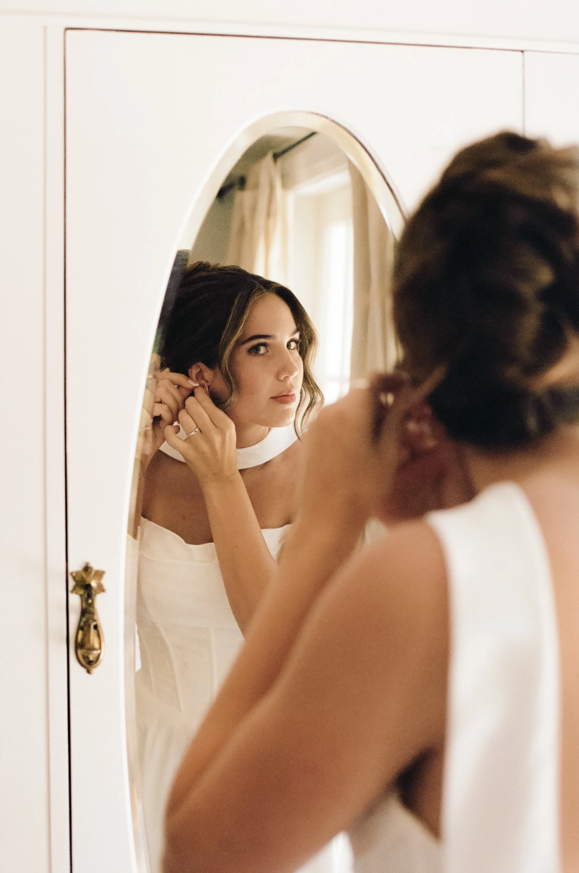 Woman getting ready, adjusting her earring while looking in a mirror, in a well-lit room.
