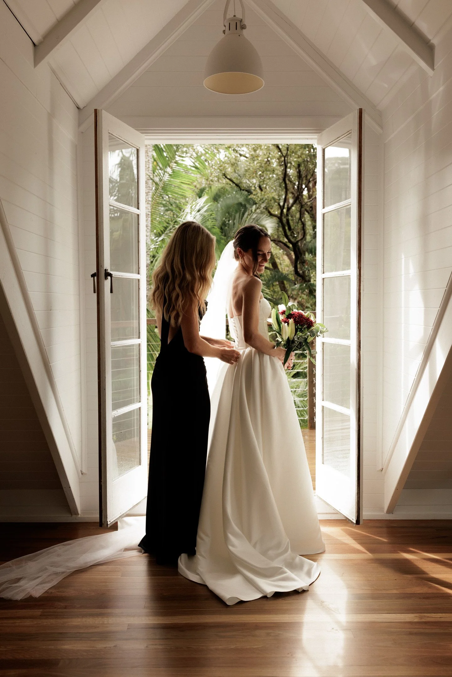 A bride in a white wedding gown holding a bouquet of flowers, standing in an open doorway with two women, one in a black dress, in a bright, indoor space with large windows and greenery outside.