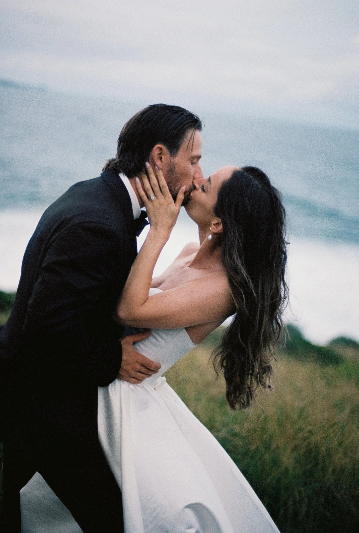 A man and woman share a kiss outdoors near the ocean, with the man in a black tuxedo and the woman in a white dress.