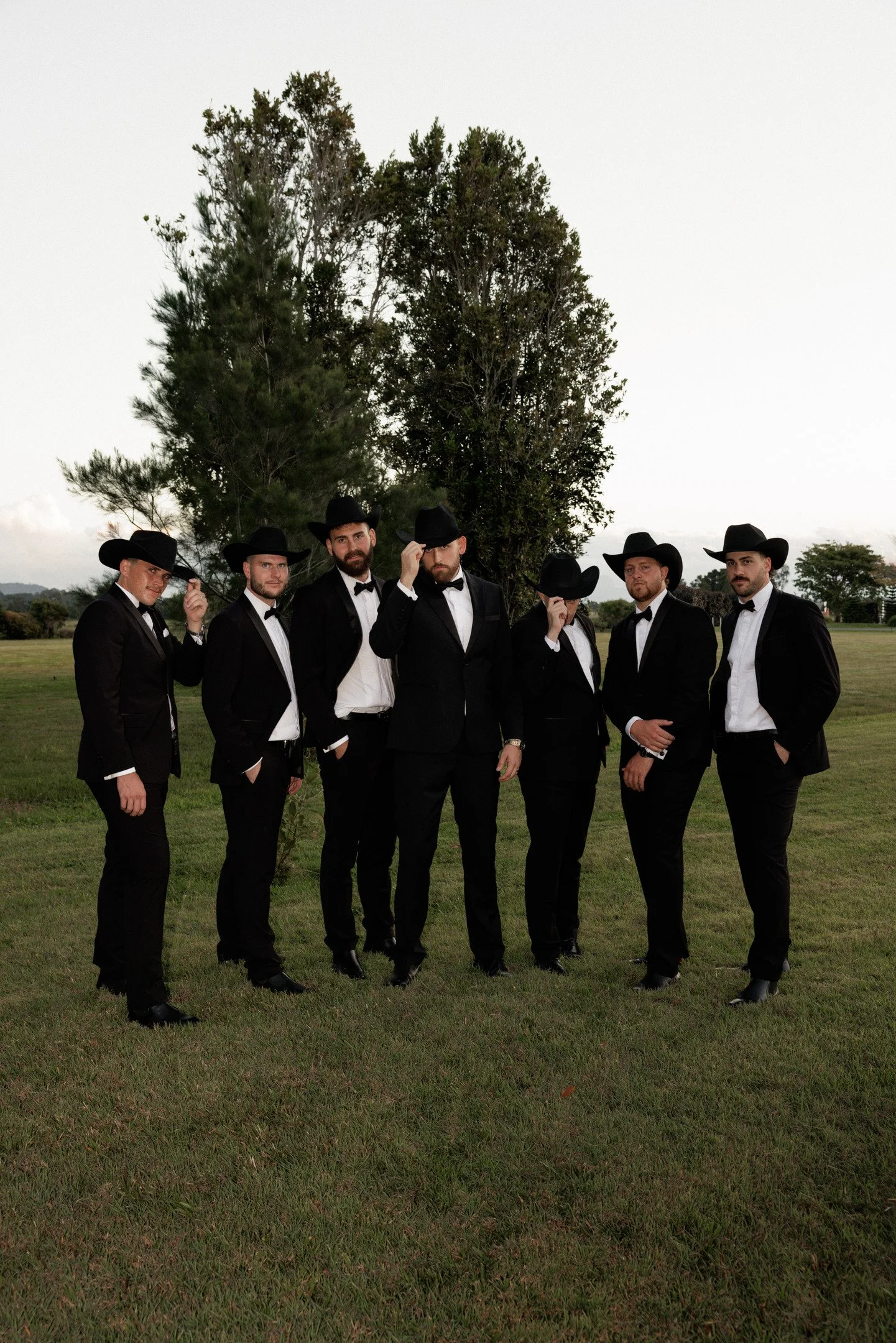 Seven men dressed in black suits, white shirts, black bow ties, and black cowboy hats standing on a grassy field in front of large trees during daytime.