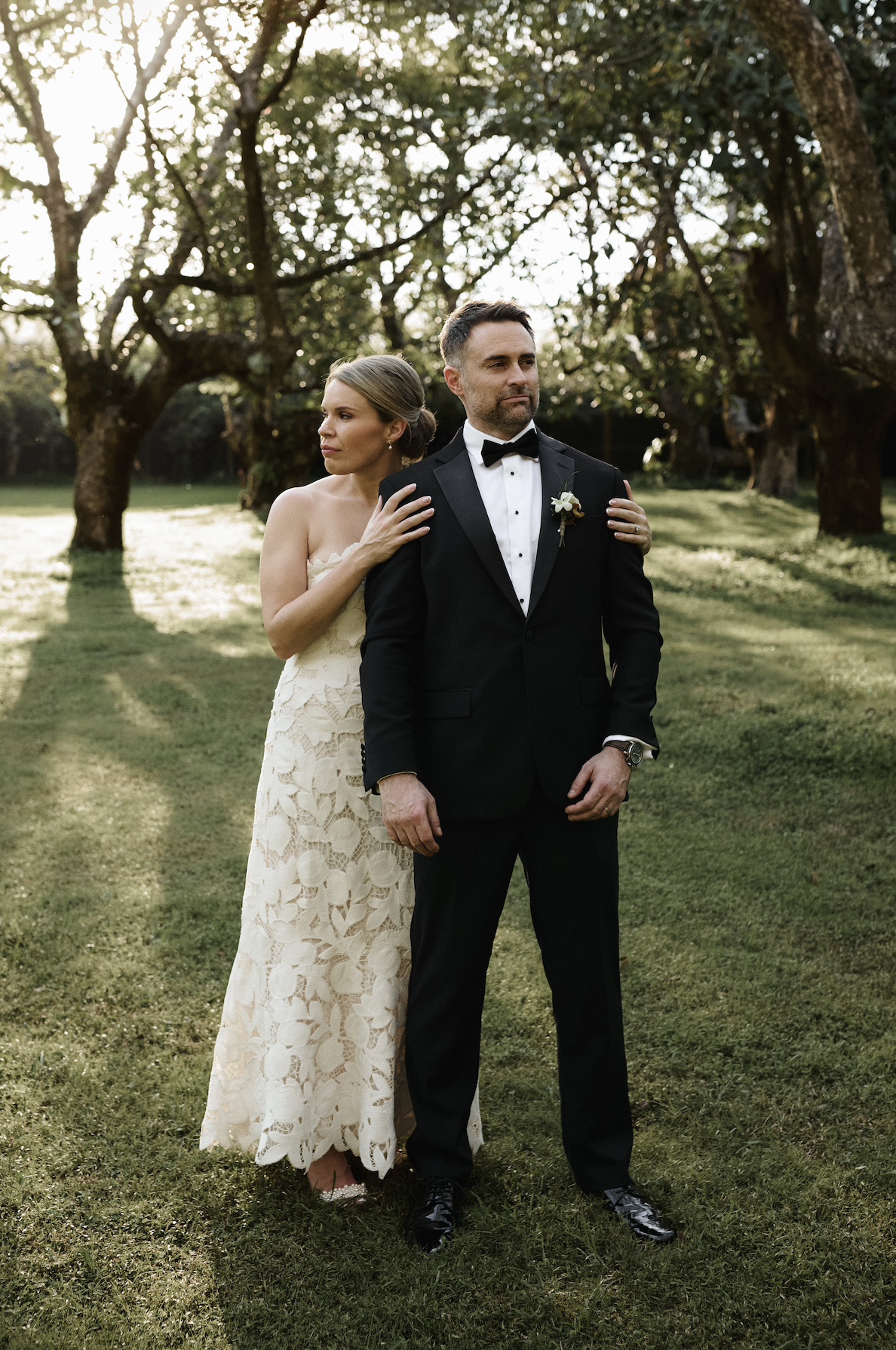 A bride in a cream lace wedding dress standing behind a groom in a black tuxedo, in an outdoor garden setting with large trees and sunlight filtering through the leaves.