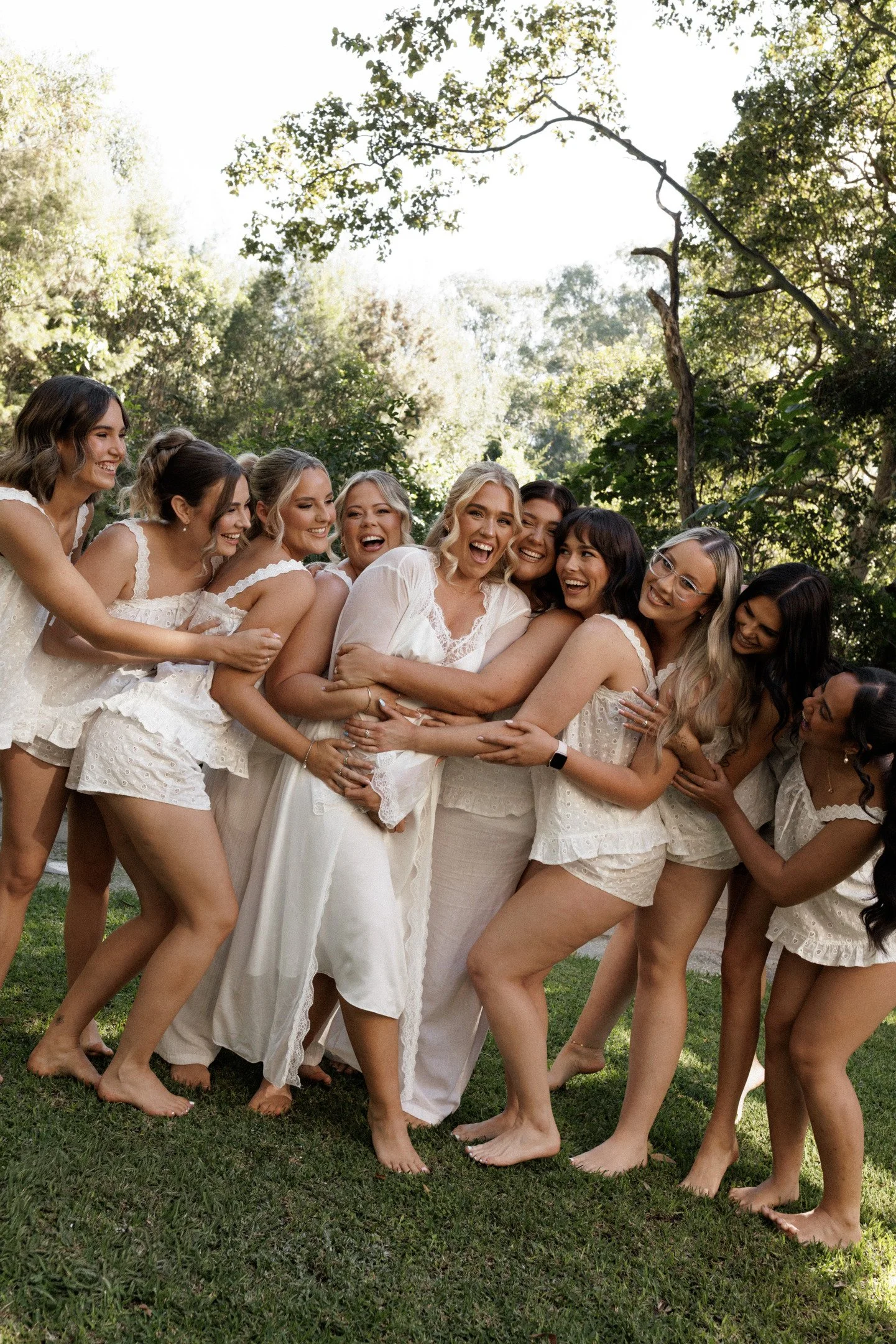 Group of women in white dresses laughing and hugging outdoors in a grassy area with trees and sunlight.