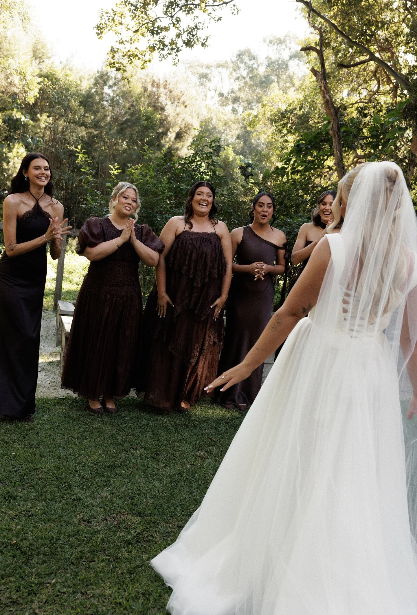 A bride in a white wedding gown with a veil stands outdoors facing five bridesmaids dressed in black and dark purple gowns, smiling in a wooded area during the daytime.