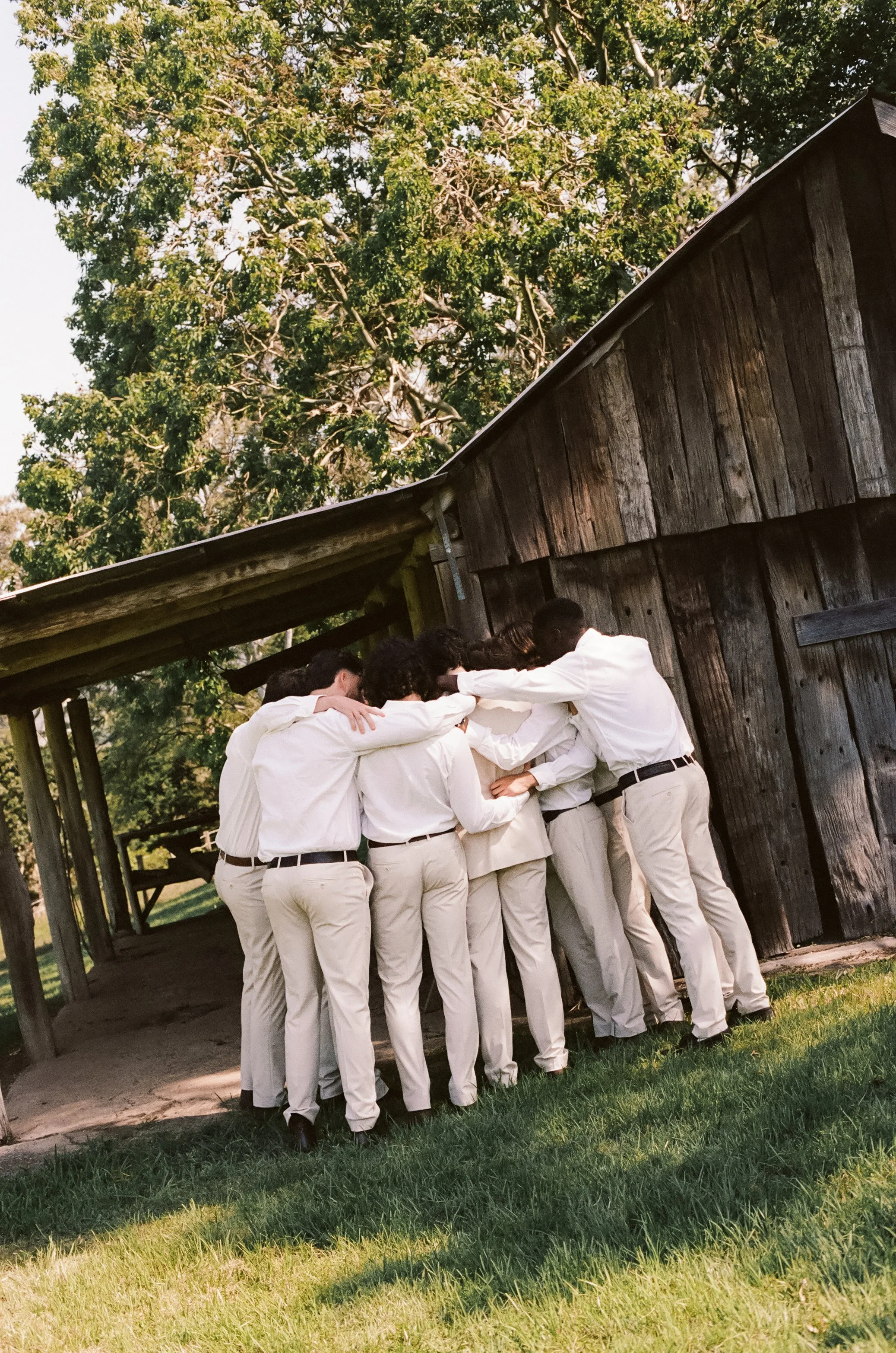 Group of people dressed in white clothing are huddled together in a circle outside a rustic wooden building with green grass and trees in the background.
