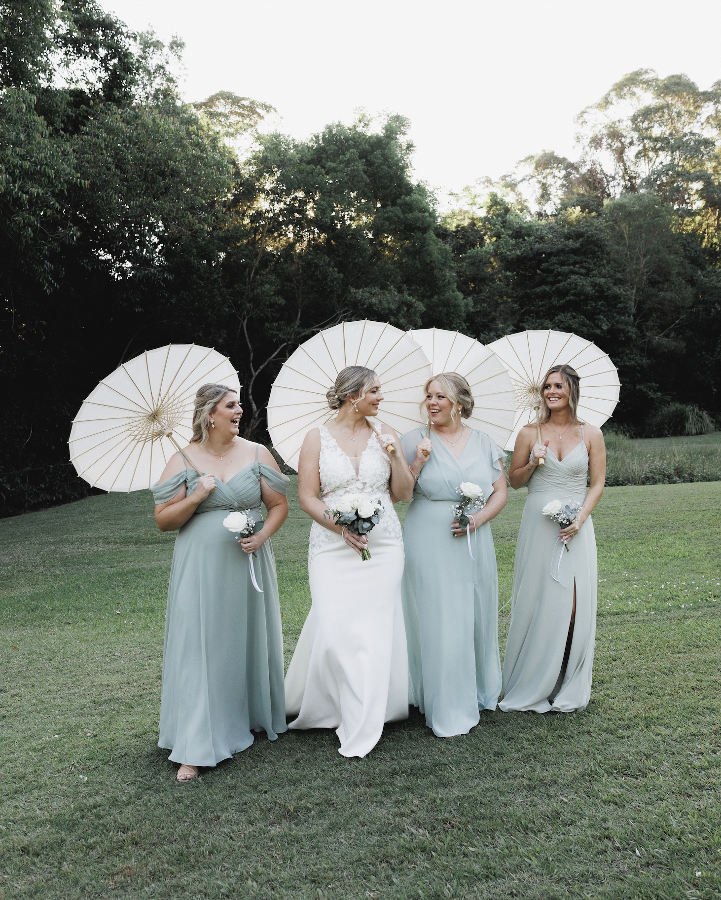 Four women, including a bride and three bridesmaids, standing outdoors on a grassy area, holding white parasols and bouquets, smiling and talking with trees in the background.