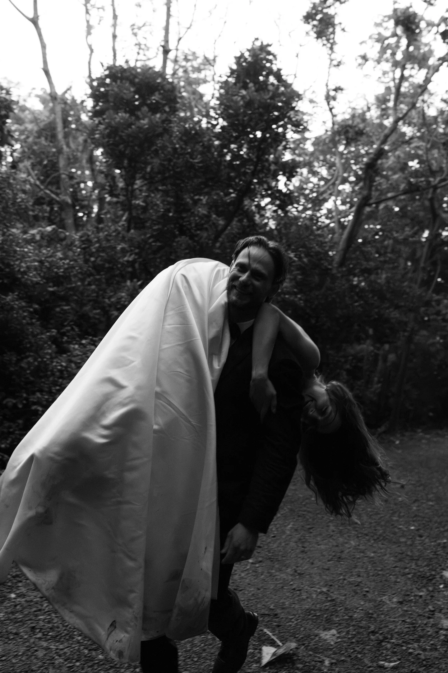 Black and white photo of a man with a beard smiling, wearing a white coat or robe, standing outdoors among trees, with a woman below him bending down.
