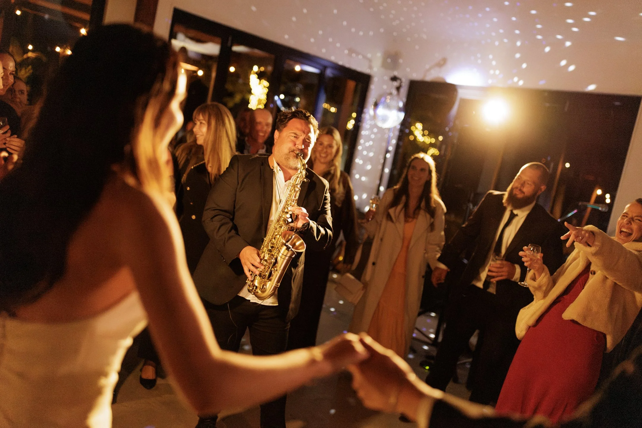A man playing the saxophone at a party with people dancing and smiling, decorated with string lights and a disco ball.