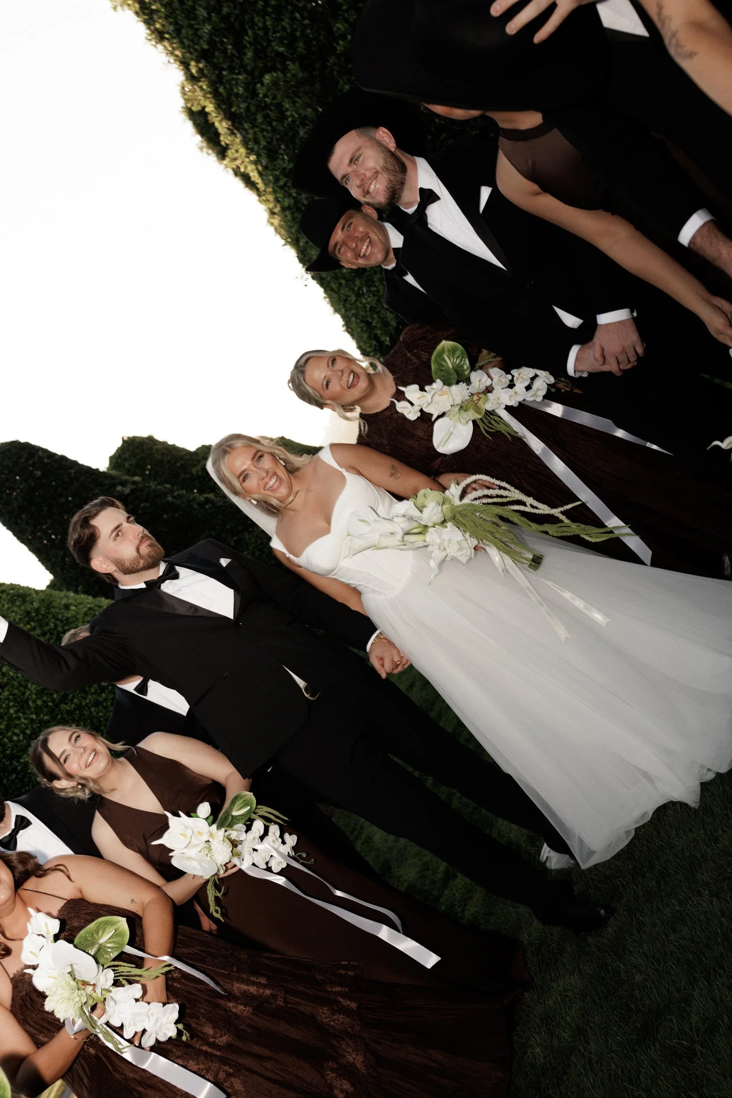 A group of people dressed in formal attire at an outdoor wedding, with women in white dresses holding floral bouquets and men in tuxedos. The background features greenery and trees.