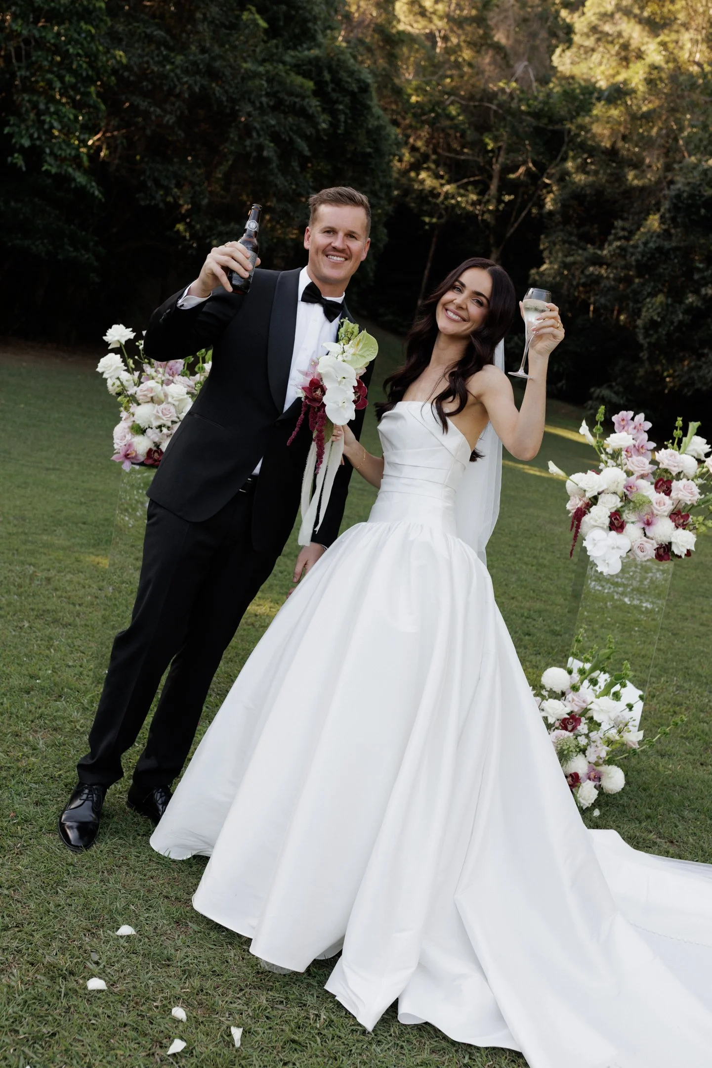 A newlywed couple dressed in wedding attire celebrating outdoors, holding glasses of champagne and smiling, with a backdrop of trees and floral arrangements.