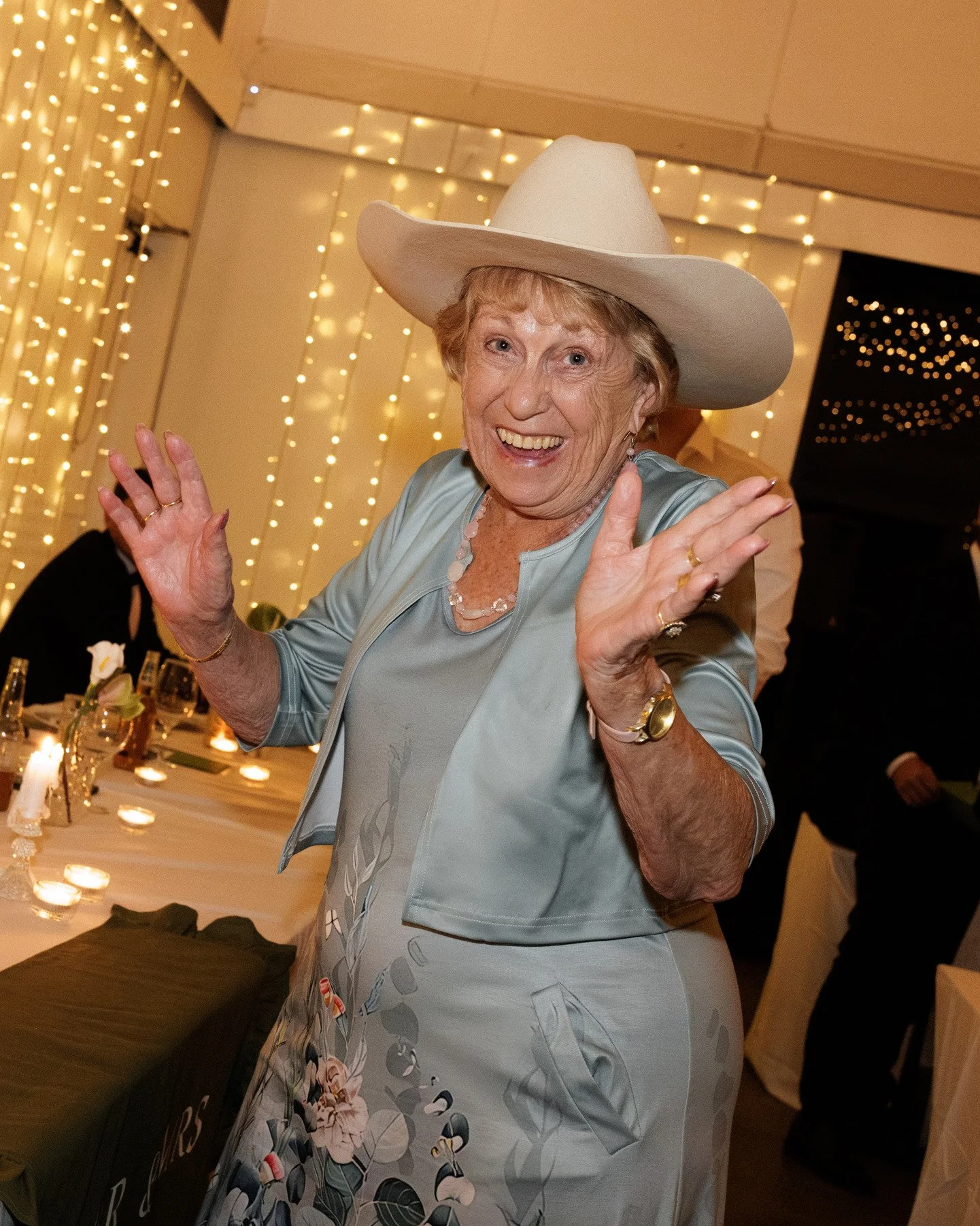 Smiling elderly woman wearing a large white hat and floral dress, dancing at a festive party with string lights in the background.
