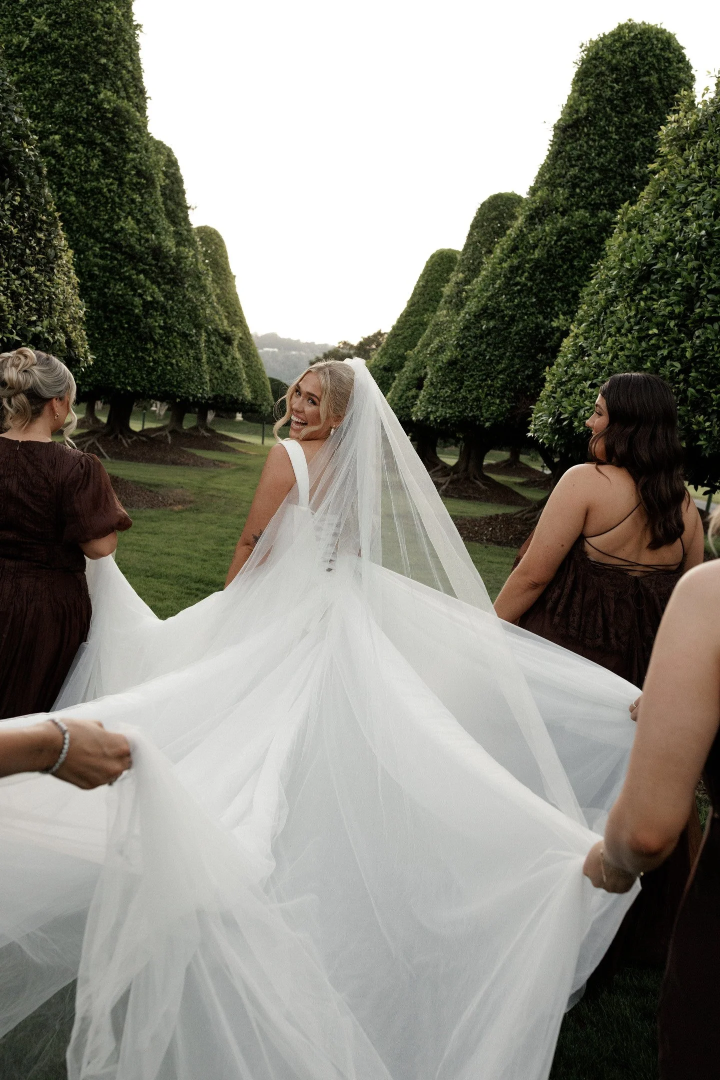 A bride in a white wedding dress with veil smiling as she walks through a garden with three women in brown dresses holding her train.