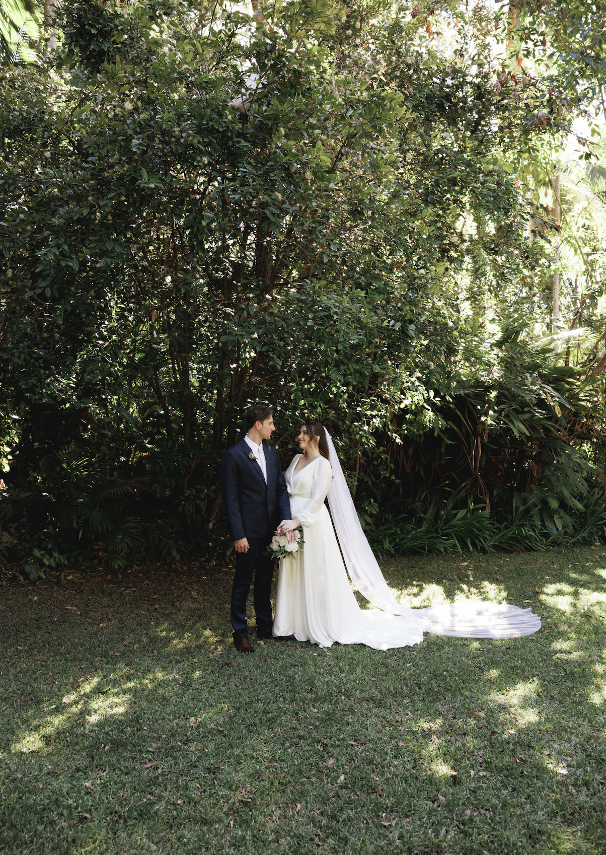 A bride and groom stand on grass in front of greenery, holding hands and looking at each other during their wedding photoshoot.
