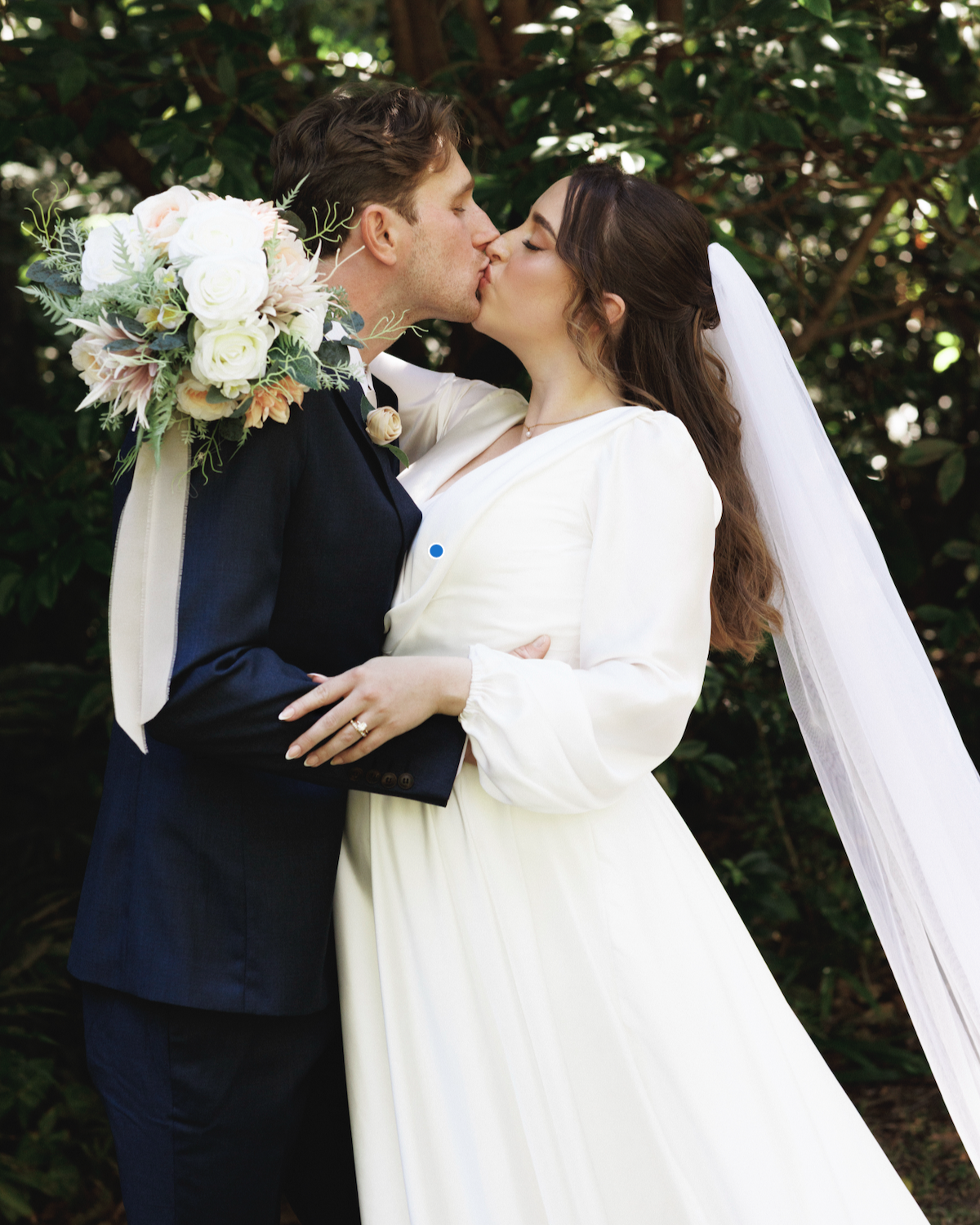 A bride and groom kissing outdoors, dressed in wedding attire, with the bride holding a bouquet of white and pastel flowers and greenery.