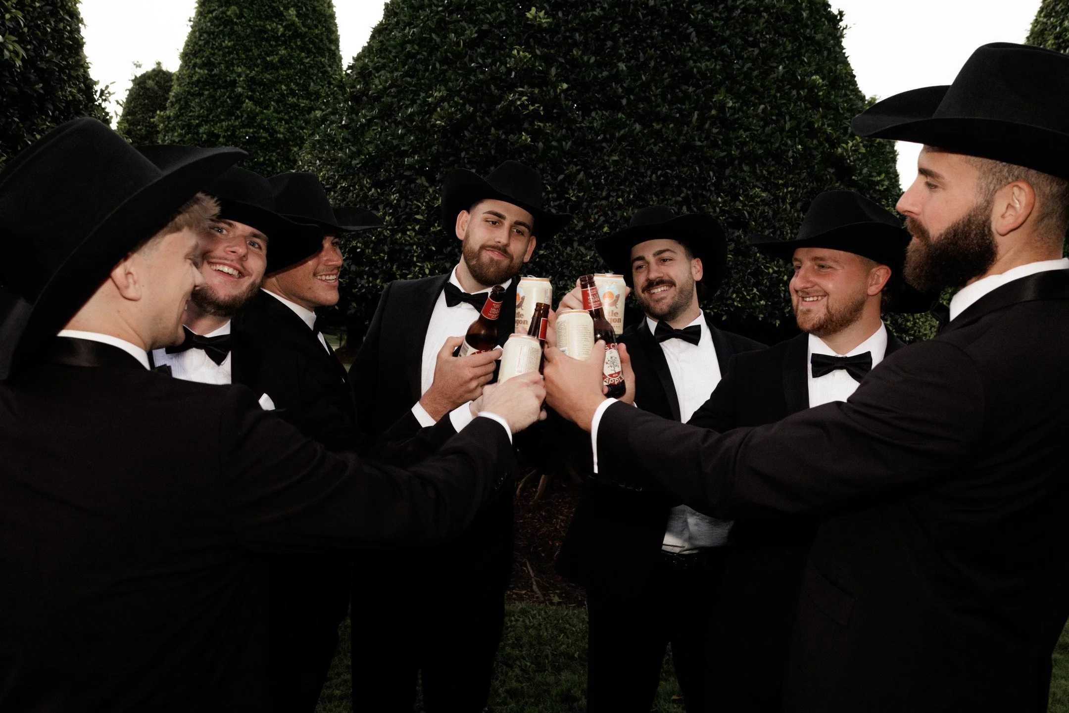 Group of men dressed in tuxedos and cowboy hats holding and clinking bottles in celebration outdoors surrounded by large, trimmed green bushes.