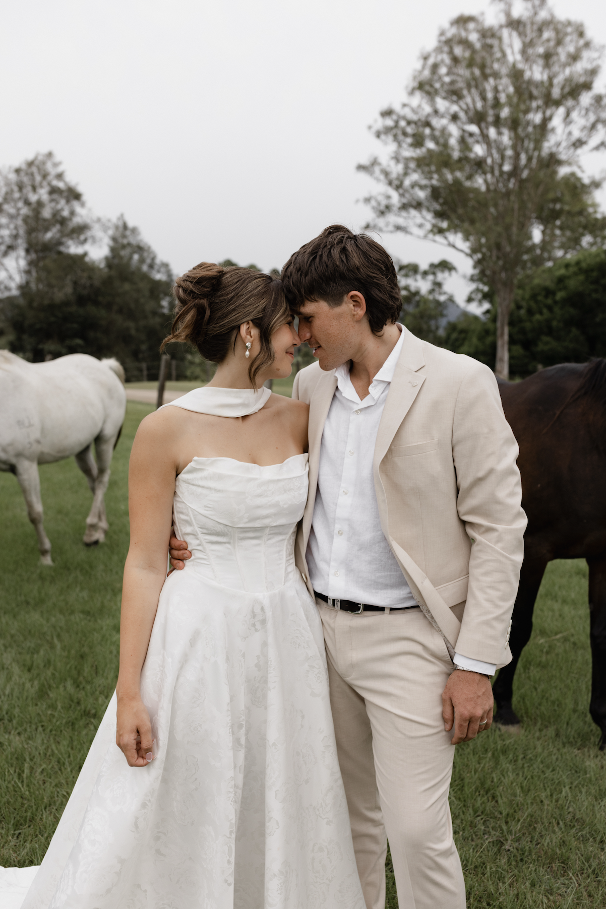 A bride and groom lean their foreheads together in a grassy field with horses and trees in the background.