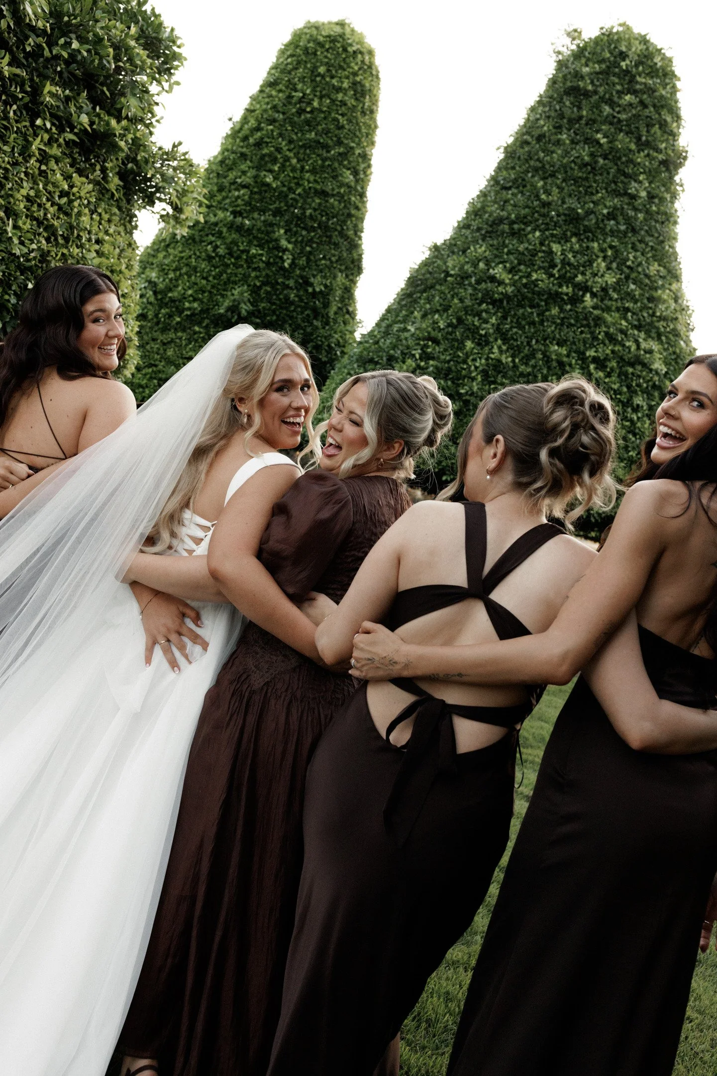 Group of women, including a bride in a white wedding dress, laughing and smiling outdoors with tall, shaped green trees in the background.