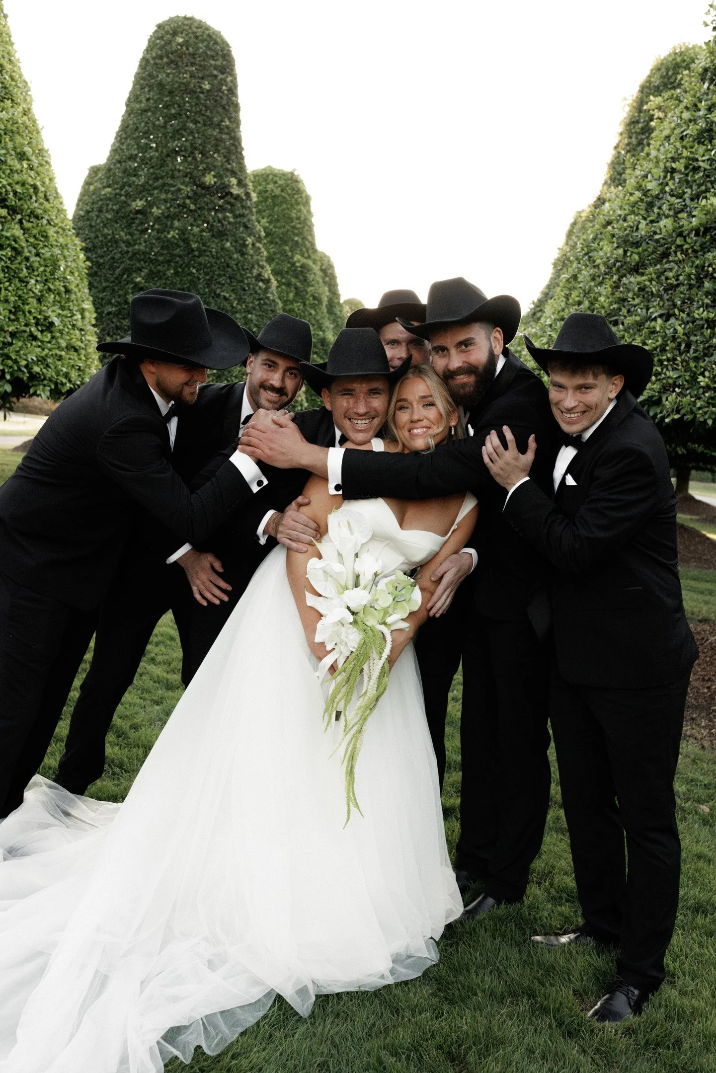 Group of six men dressed in black tuxedos and wide-brimmed hats surrounding and embracing a smiling bride in a white wedding gown holding a bouquet of white flowers outdoors on green grass with large, trimmed bushes in the background.
