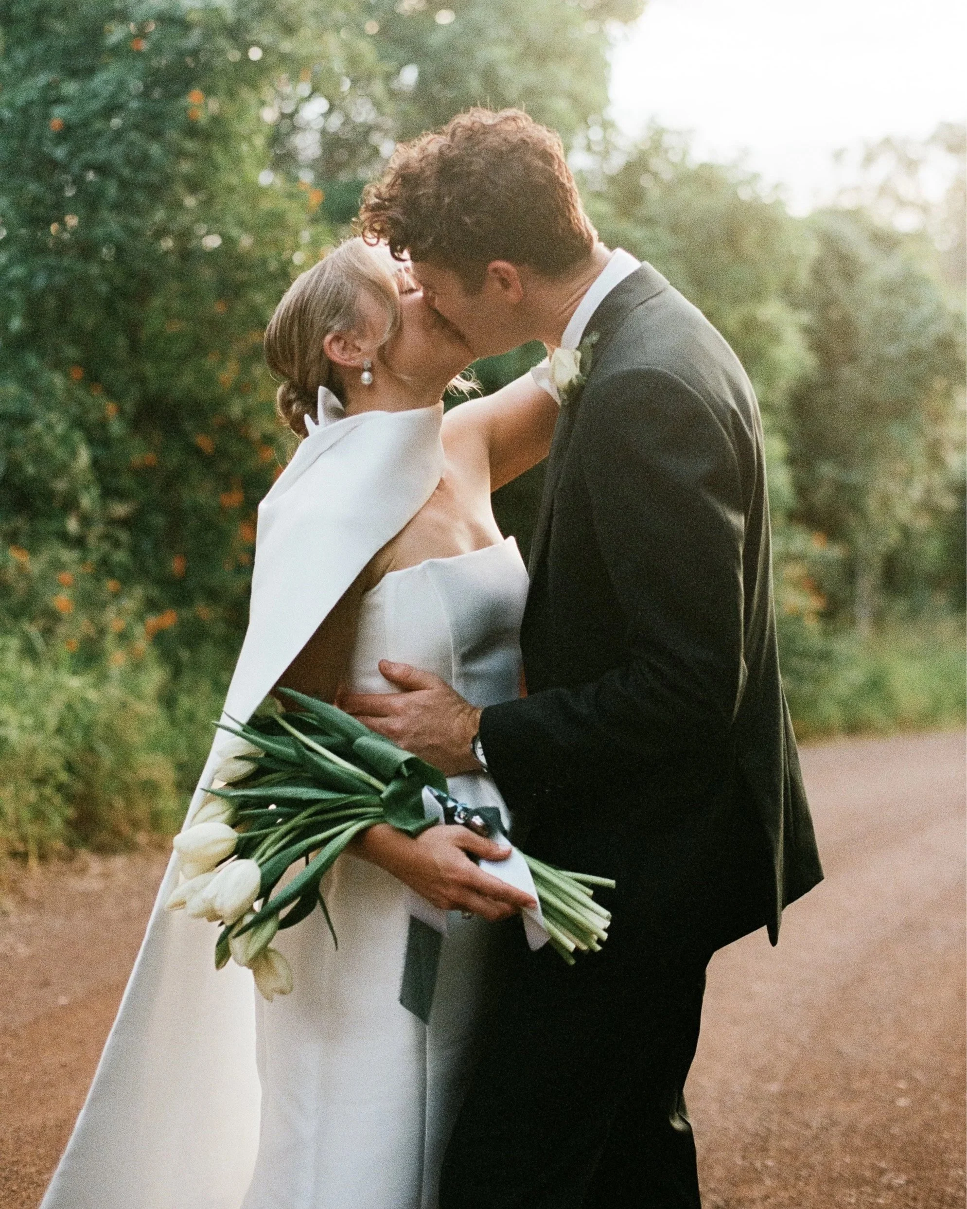 A bride and groom kiss outdoors during their wedding, with trees in the background. The bride holds a bouquet of white tulips, and both are dressed in formal wedding attire.