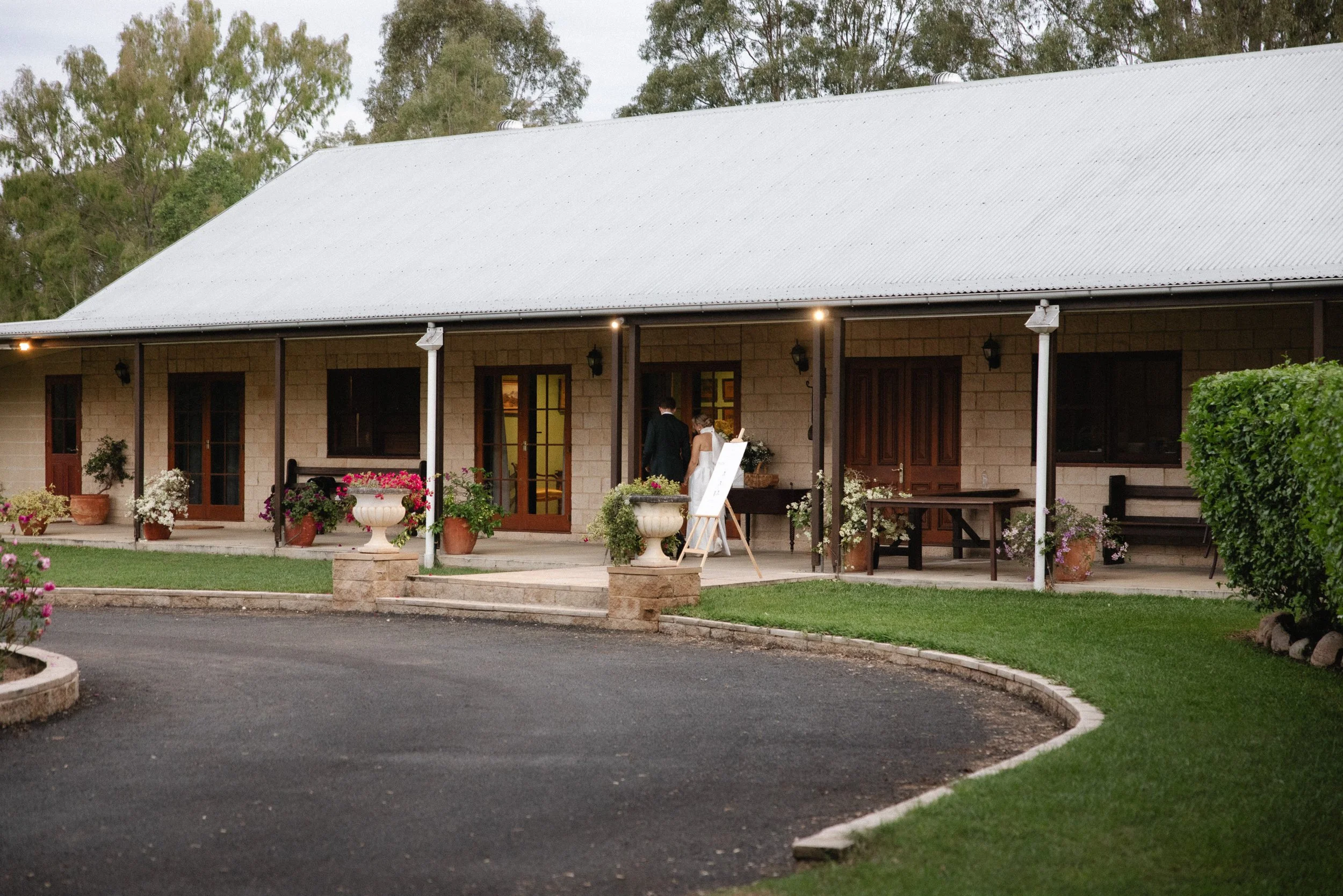 Rustic building with a tin roof, featuring wooden doors and windows, surrounded by plants and shrubs in a well-maintained garden area. A couple is entering through one of the doors - Elevated Weddings