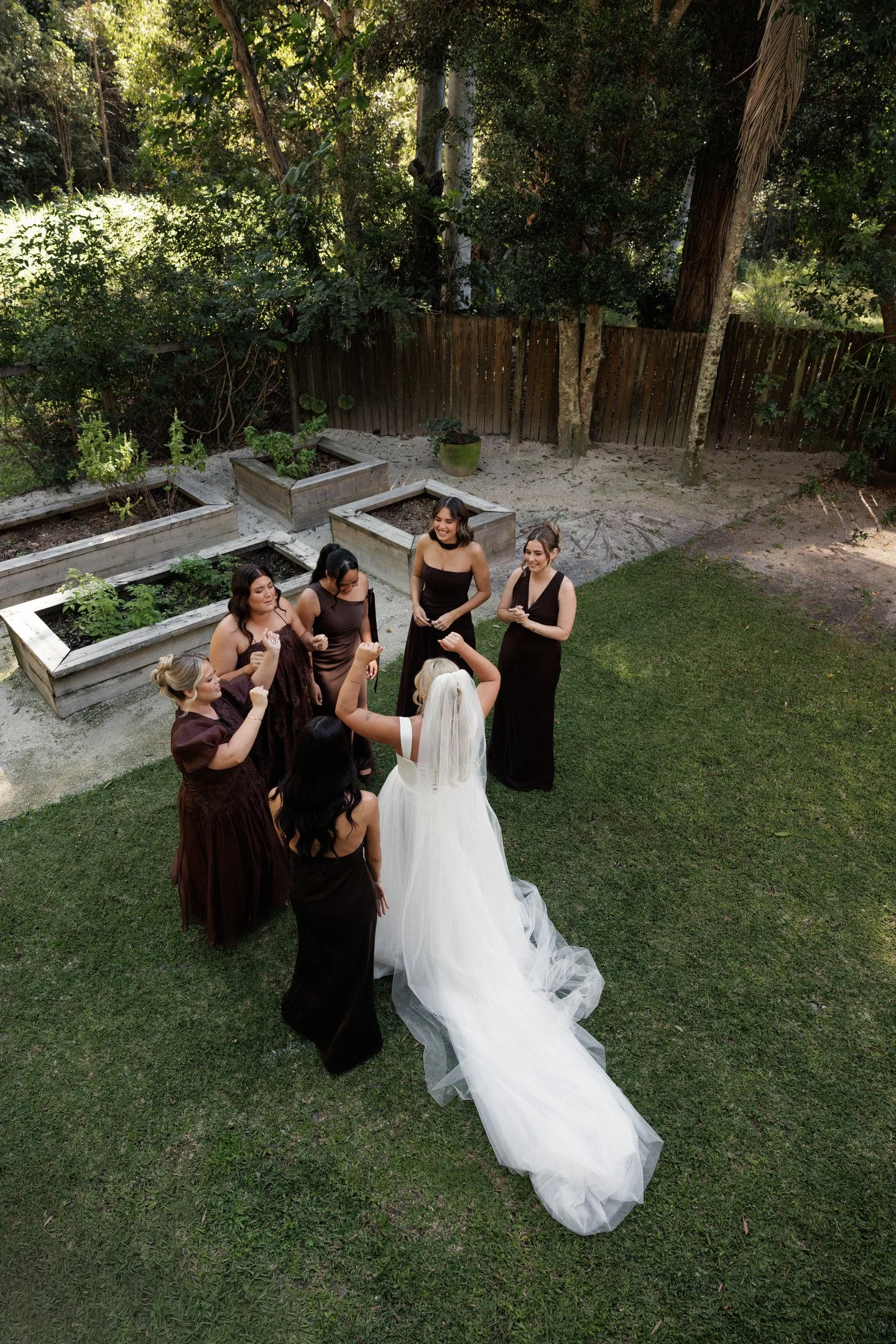 A group of women gathered outdoors, some in matching dark dresses, surrounding a bride in a white wedding gown with a long train. They are smiling and appear to be celebrating.