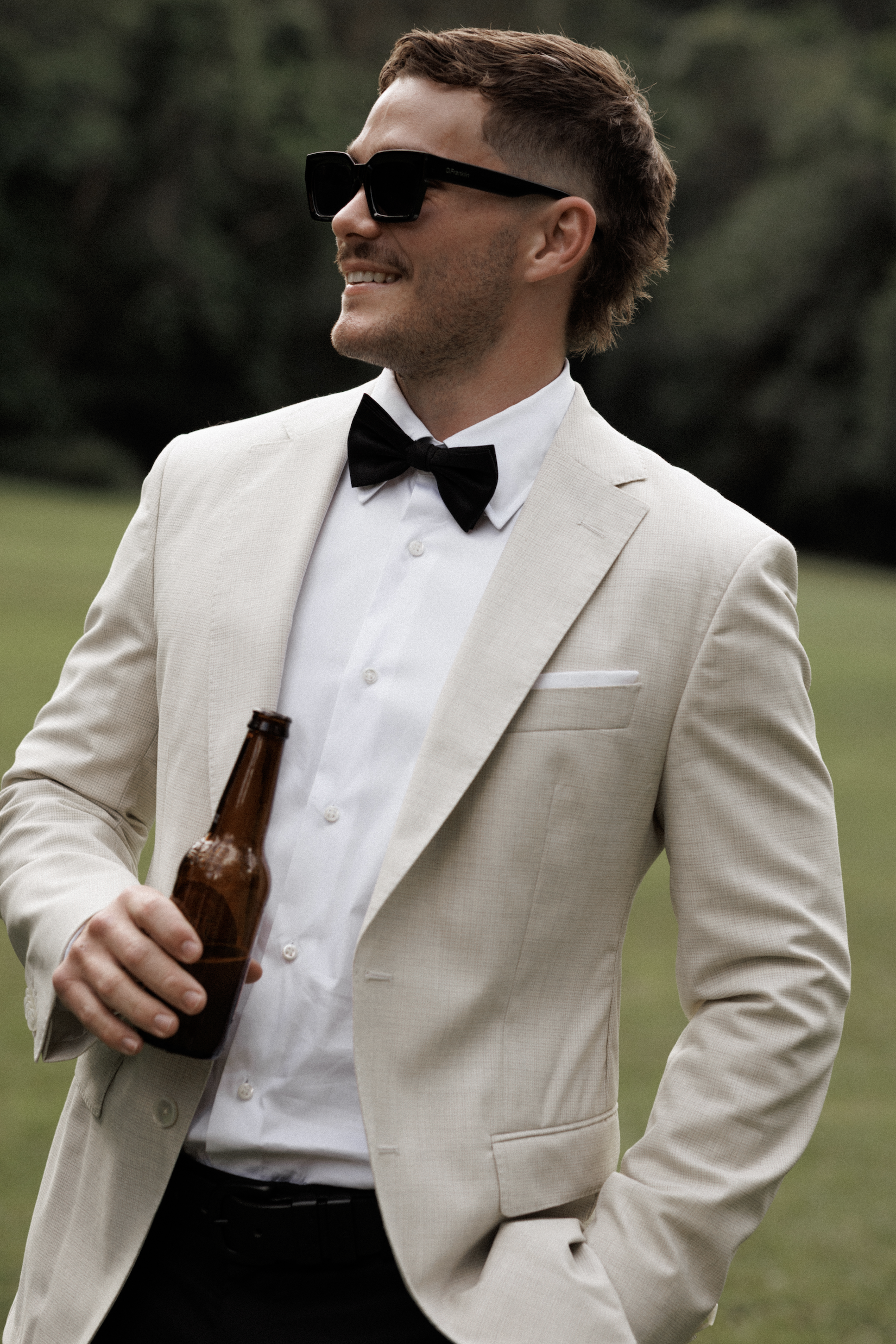 A man dressed in a light-colored suit, white shirt, and black bow tie, wearing sunglasses, holding a beer bottle, standing outdoors with a blurred green background.