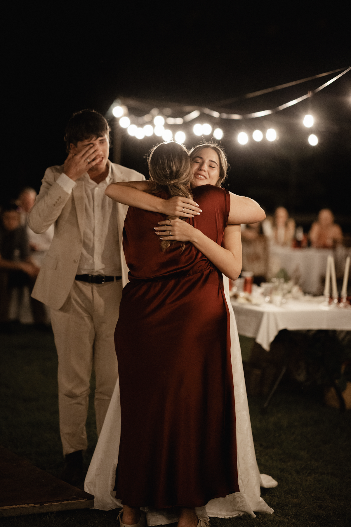 Three people hugging and emotional at an outdoor nighttime celebration, with string lights overhead and tables set in the background.