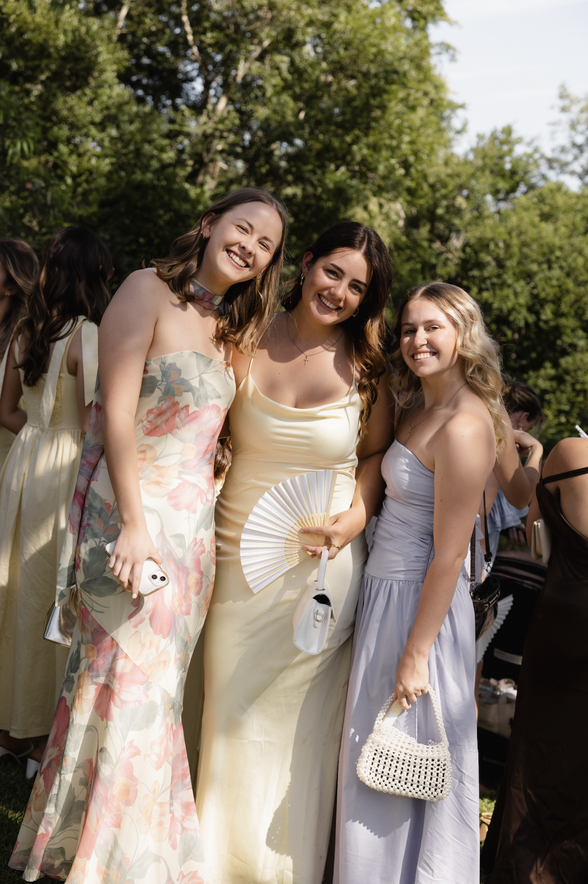 Three young women in pastel dresses smiling outdoors, holding fan, purse, and phone at a gathering with greenery in the background.