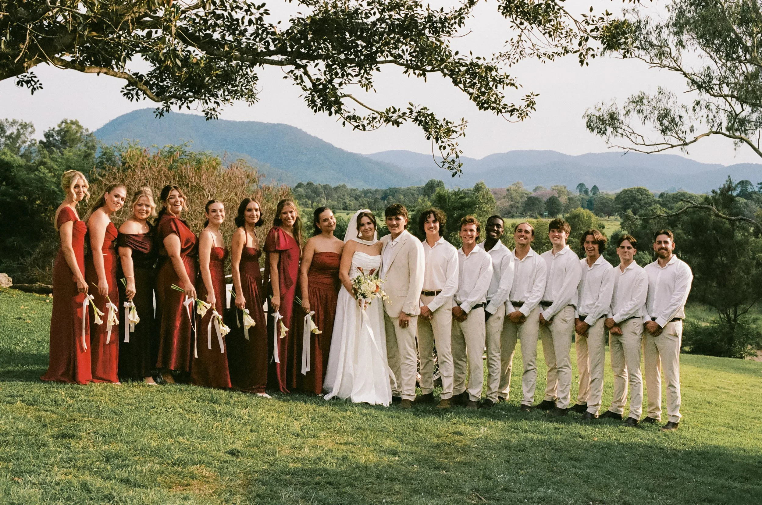 A wedding party outdoors with the bride, groom, bridesmaids in red and groomsmen in white, standing on grass with trees and mountains in the background.