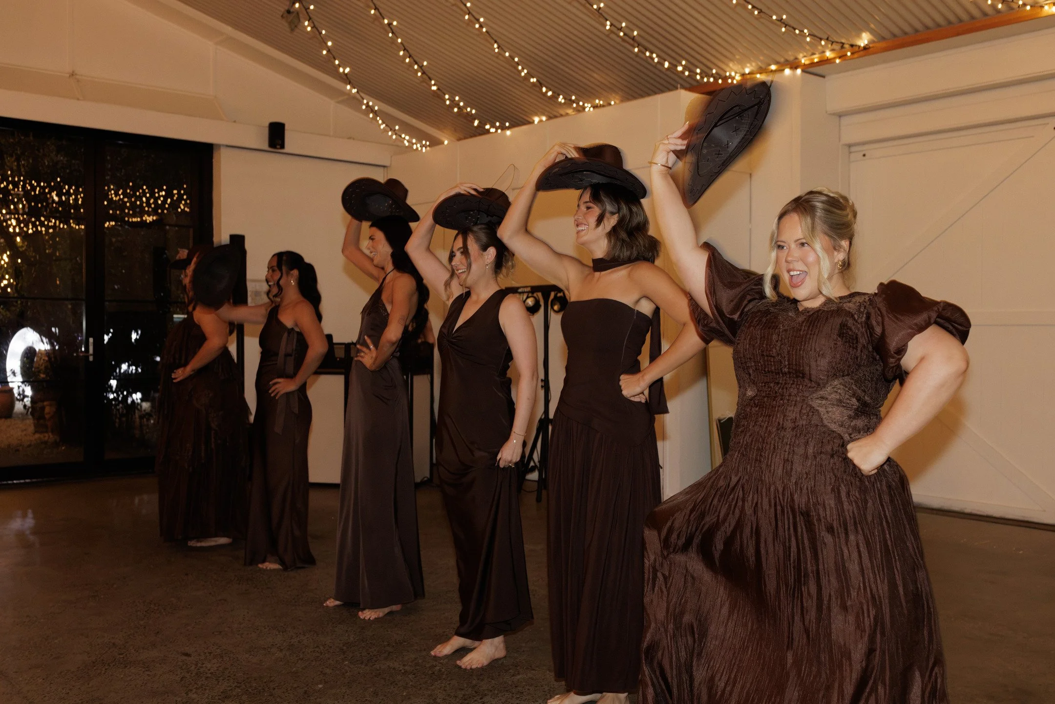 Group of women in black dresses performing a choreographed dance with cowboy hats in a decorated indoor event space.