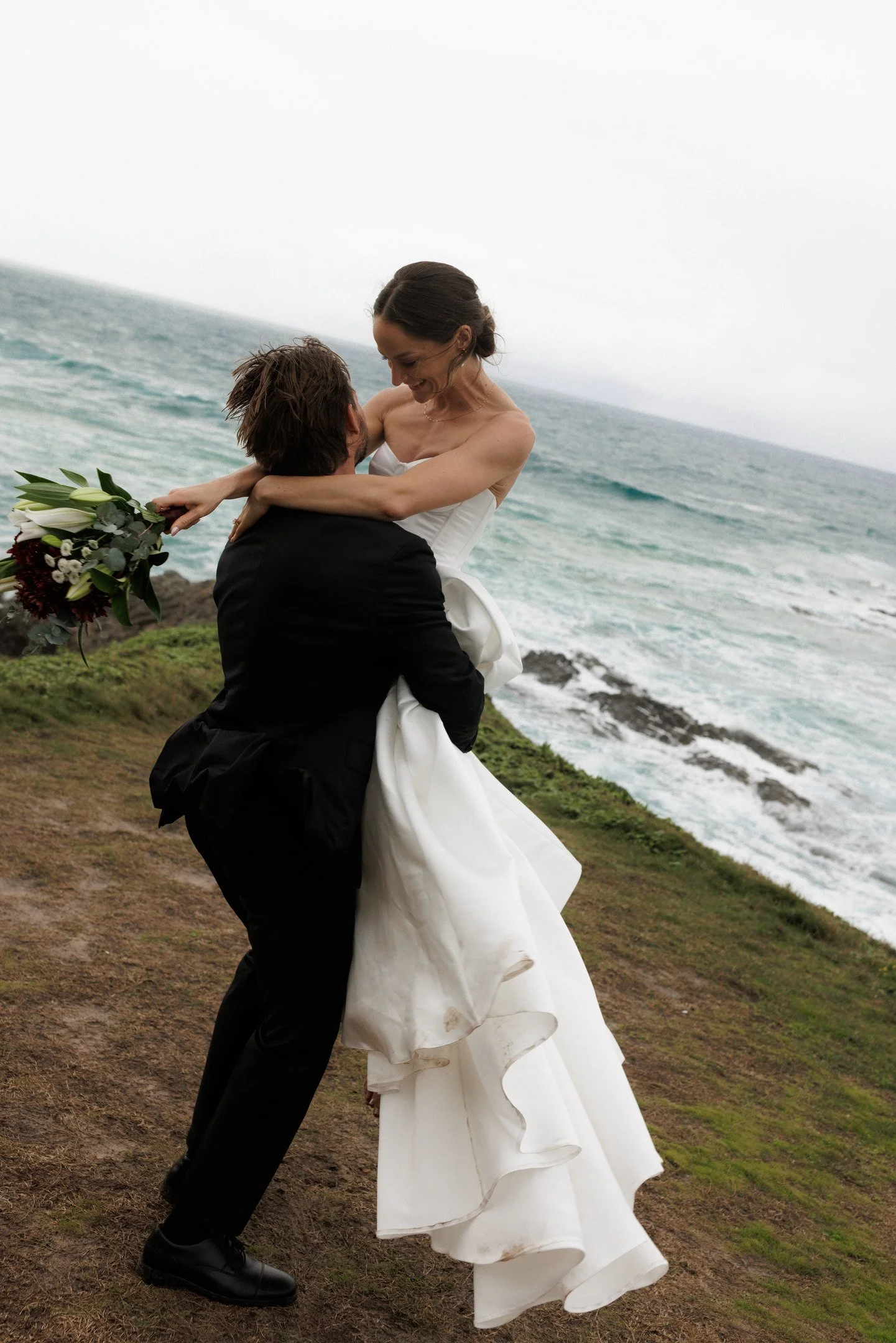 A couple, dressed in wedding attire, embracing on a grassy cliffside with the ocean in the background, celebrating their wedding day.