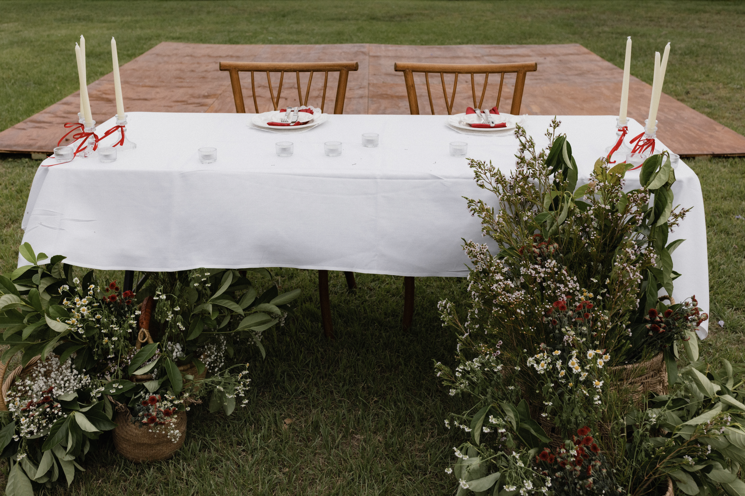 An outdoor dining setup with a table covered in white tablecloth, surrounded by plants and flowers, with a raised wooden platform in the background.