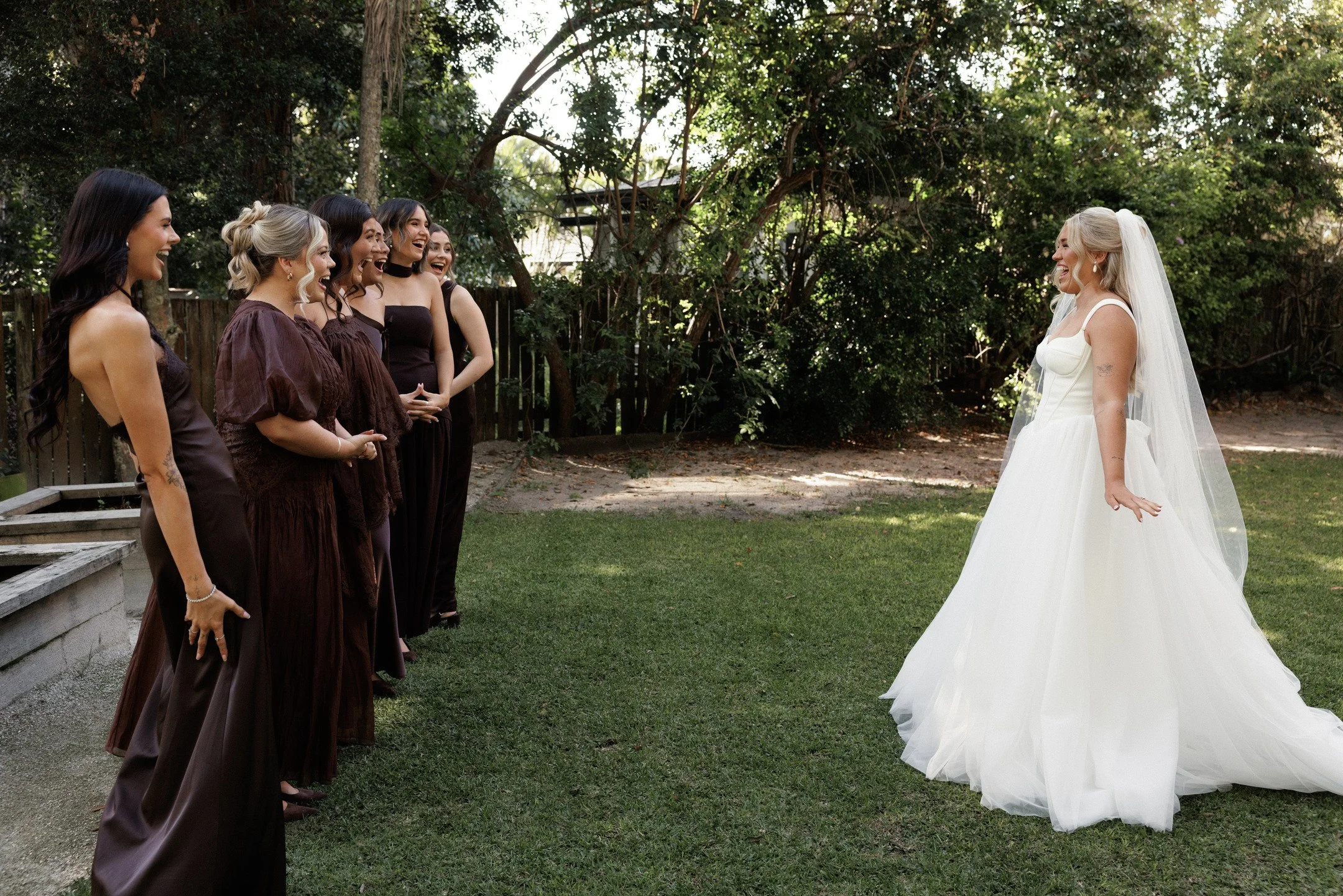 A bride in a white wedding gown with a long train and veil standing outside, smiling at five women in dark dresses who are laughing and facing her.