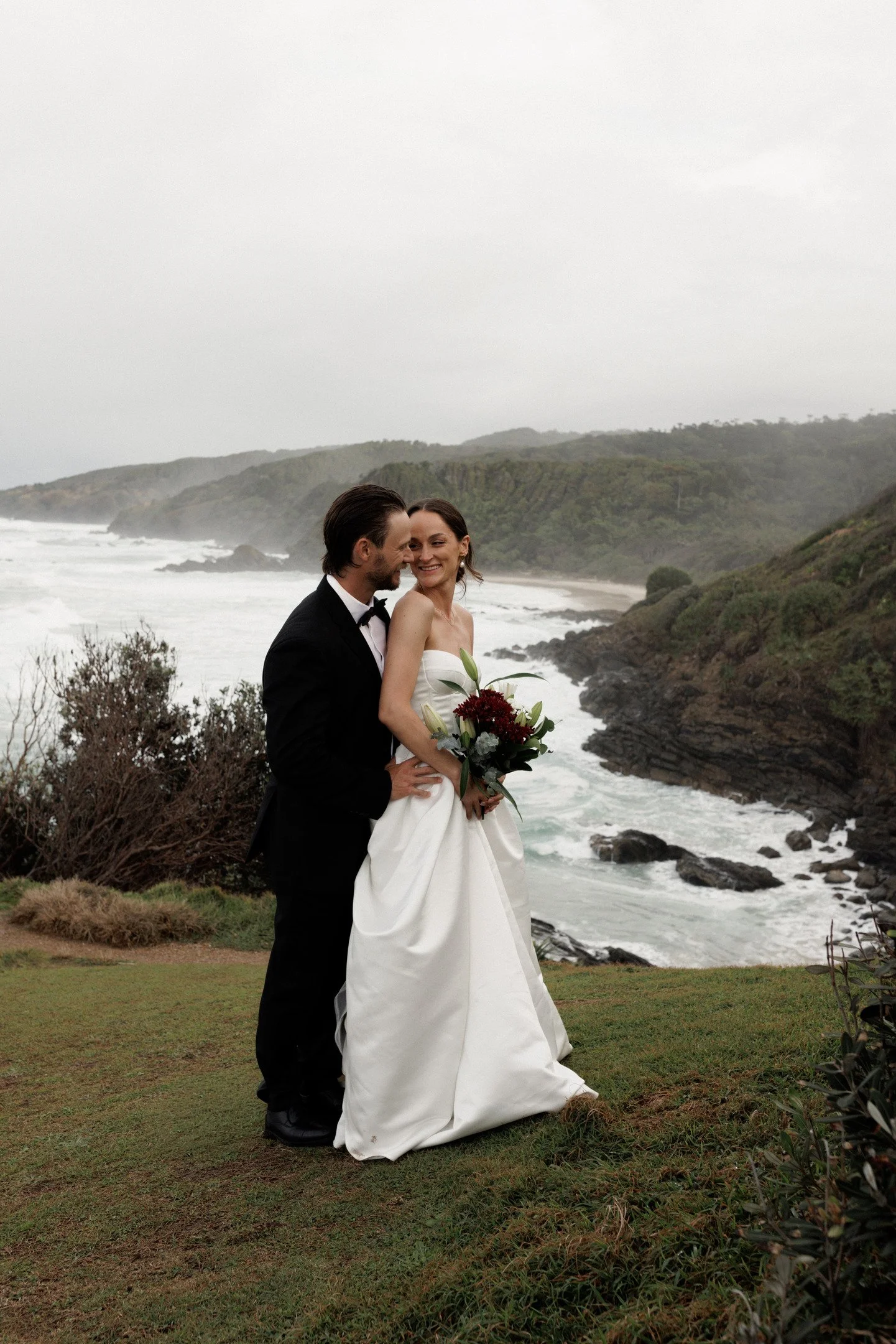 A couple in wedding attire standing on a grassy area by the ocean, with cliffs and waves in the background.