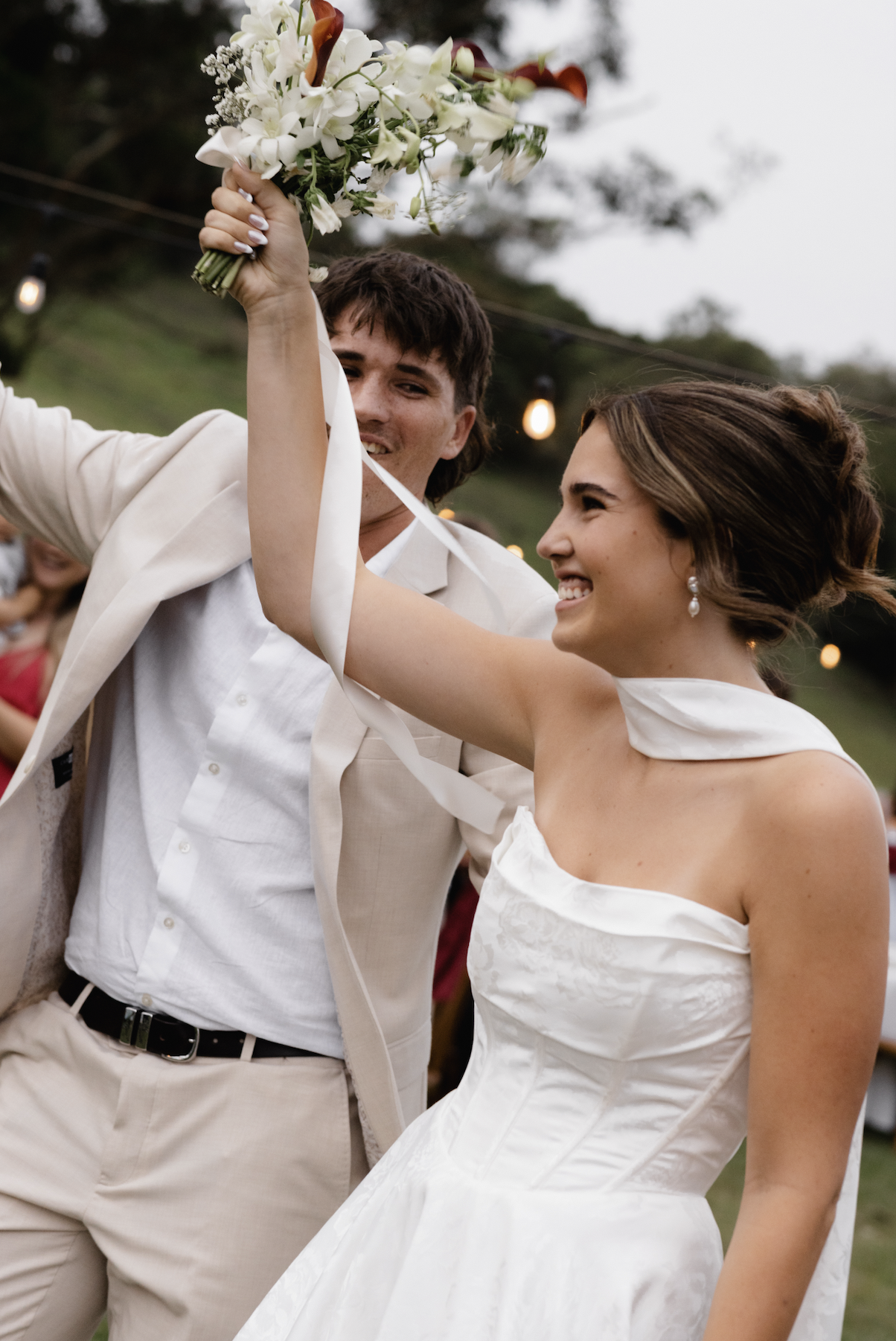 A bride and groom celebrate outdoors at their wedding, with the bride raising a bouquet of white flowers and the groom smiling behind her.