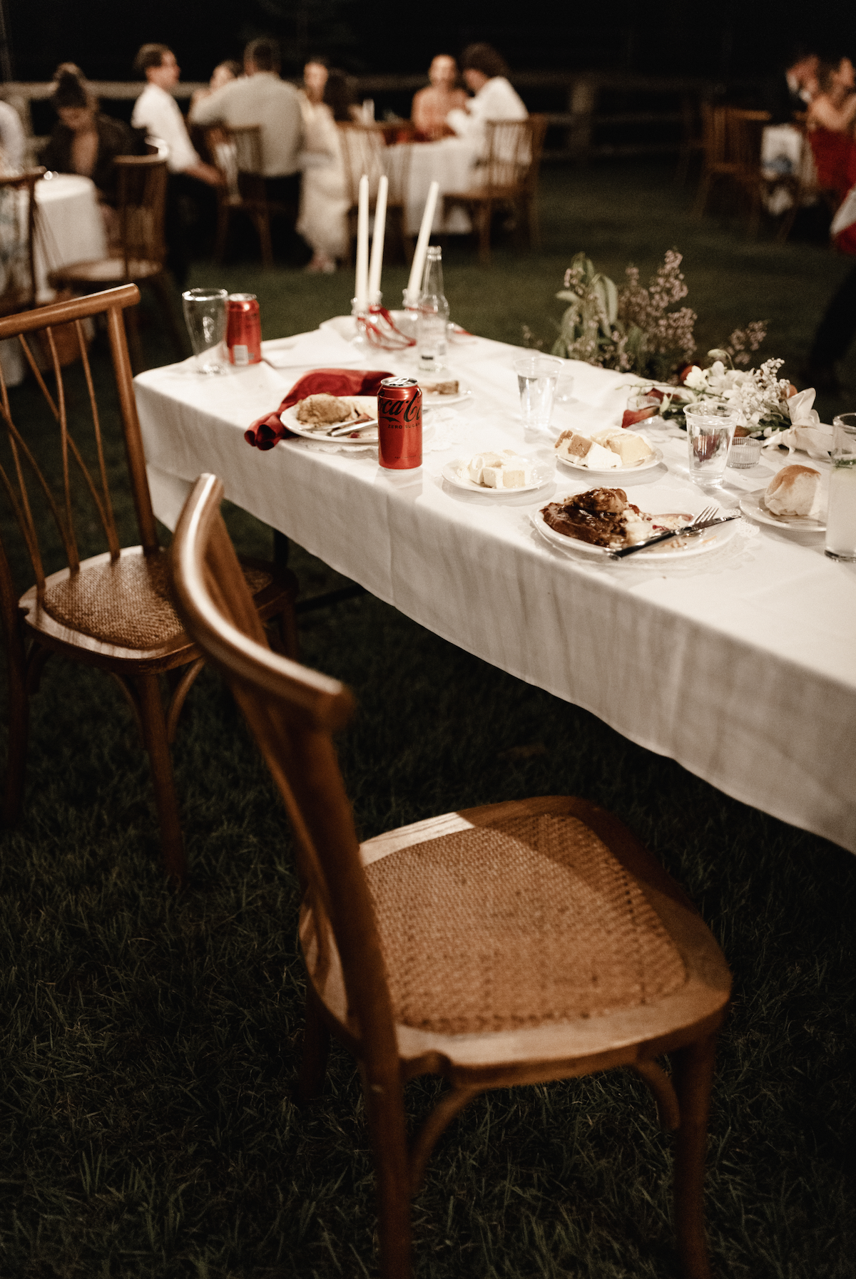 Empty chairs around a decorated dining table at an outdoor gathering, with plates, cans of Coke, and leftover food.