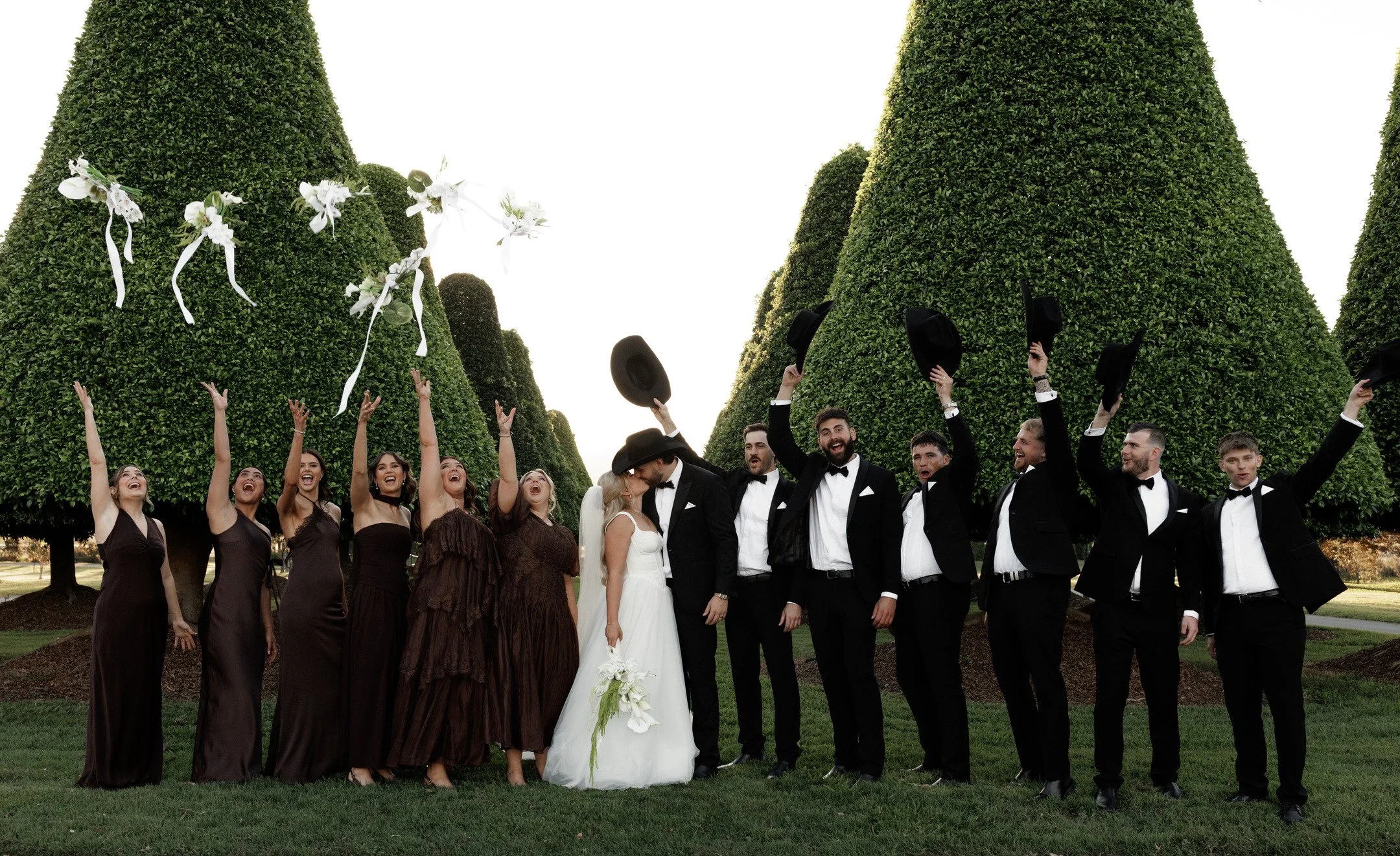 A group of people at a wedding outdoors, with large, neatly trimmed greenery in the background. The bridesmaids are dressed in dark brown gowns, and the groomsmen in black tuxedos. The bride and groom are in the center, kissing, with the groom in a tuxedo and the bride in a white wedding dress. The guests are celebrating, some throwing hats in the air, against a clear sky.