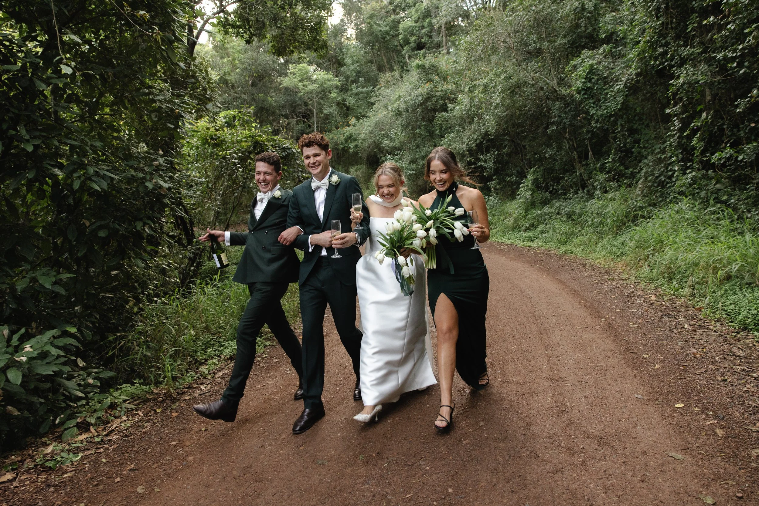 A bride and groom walking on a forest path with two friends, all smiling and holding glasses and a bottle -Elevated Weddings
