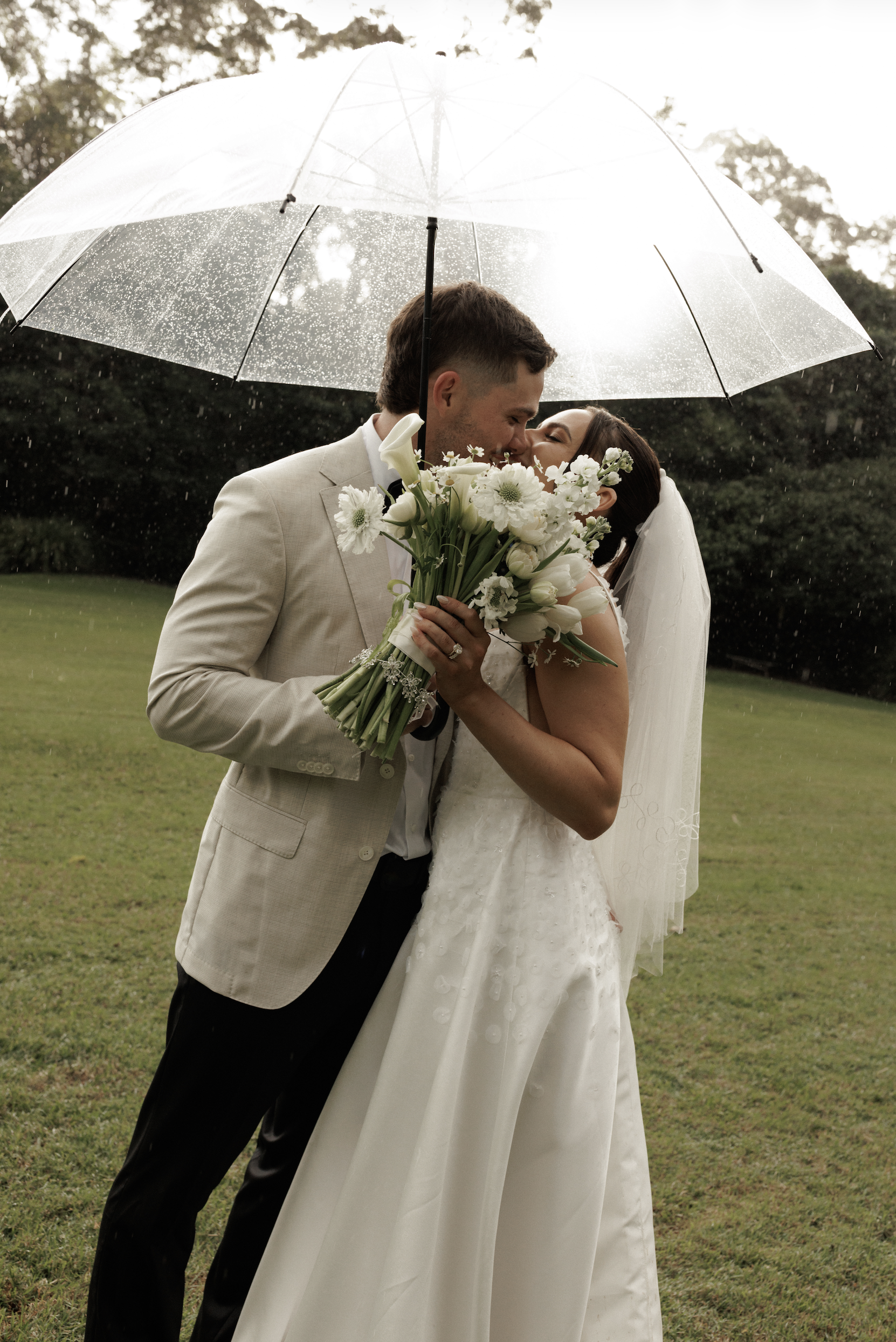 A bride and groom sharing a kiss under a transparent umbrella on a rainy day, outdoors on a grassy field with trees in the background. The bride is holding a bouquet of white flowers.