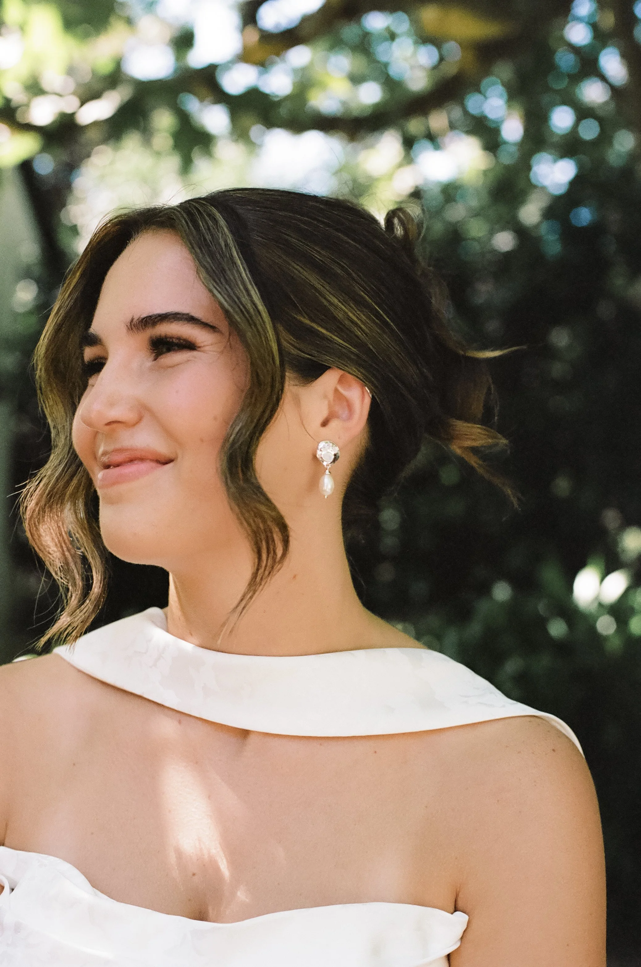 A woman with wavy brown hair and earrings, wearing a white off-the-shoulder dress, standing outdoors with a blurry green background, smiling softly.