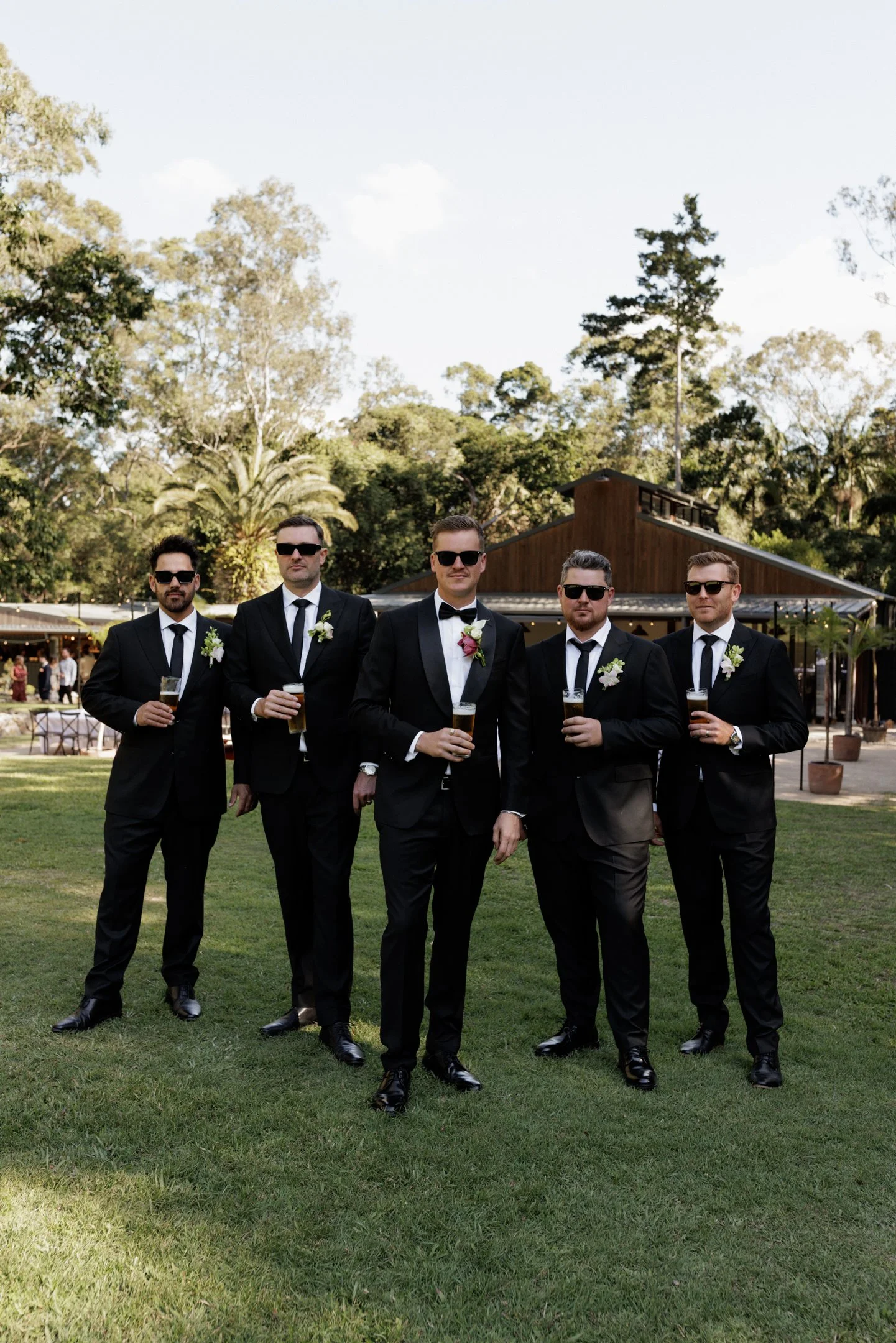 Five men in tuxedos with sunglasses, holding beers, standing on a grassy lawn at an outdoor event with trees and a wooden building in the background.