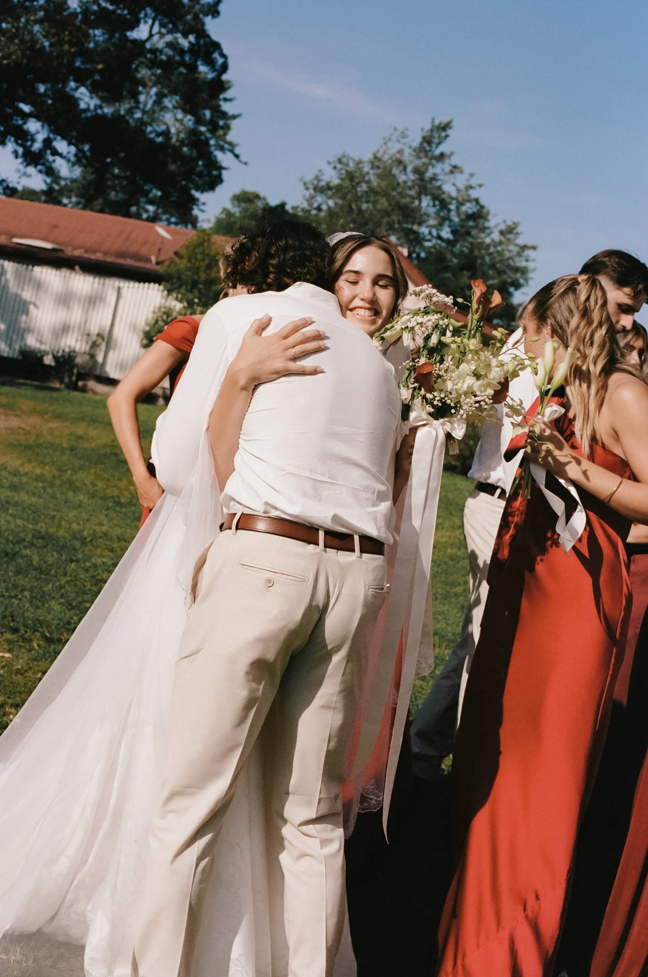 People hugging at an outdoor event, with a woman in a white dress holding a bouquet of flowers.