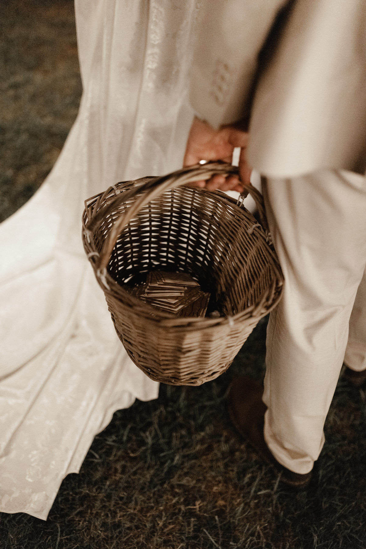 Person holding a wicker basket filled with paper slips, dressed in beige wedding attire, standing on a dark carpeted floor.
