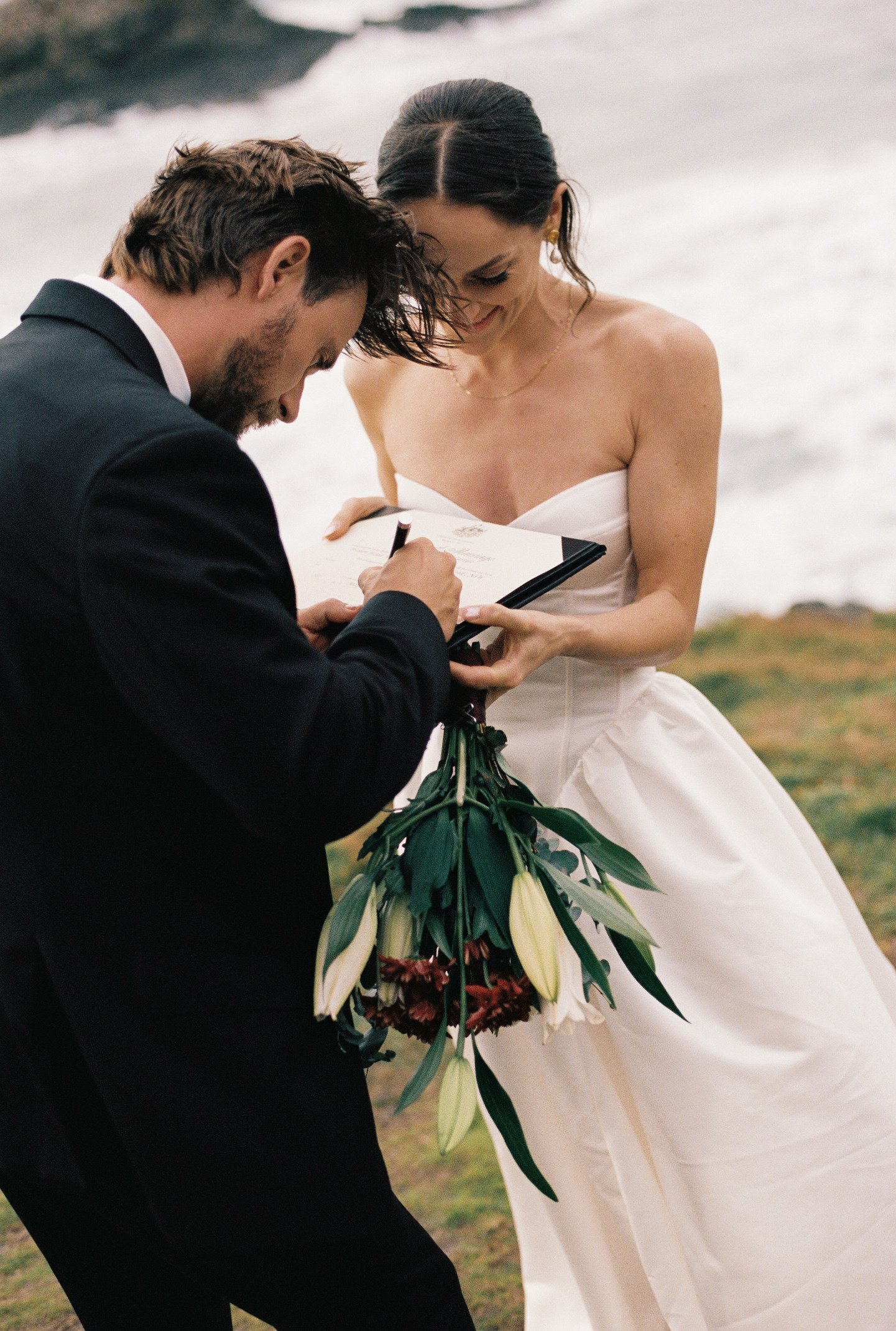 A couple getting married outdoors by the water, with the groom signing a document and the bride holding a bouquet of flowers, both smiling.