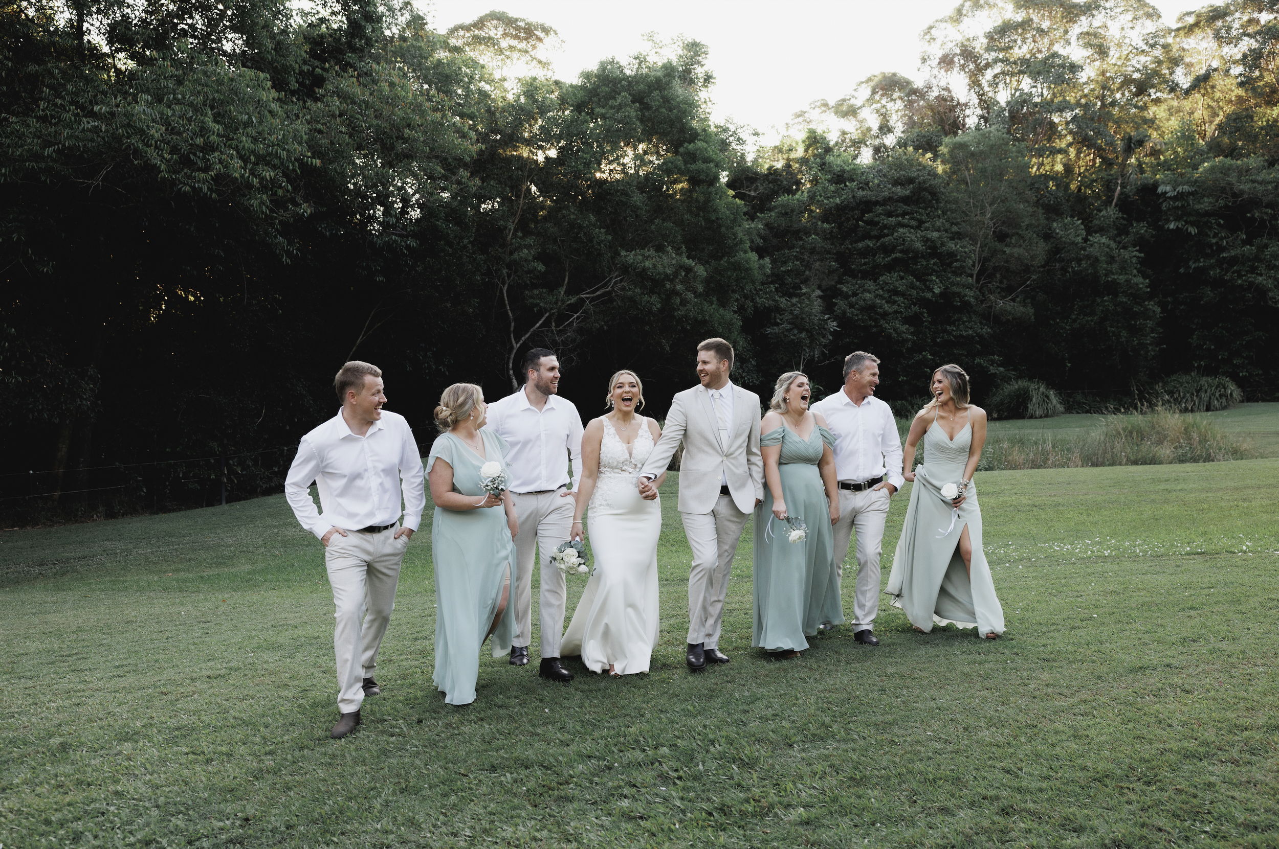 Wedding party of nine people, including brides and grooms, walking on grass in an outdoor green park, smiling and holding flowers, with trees in the background.