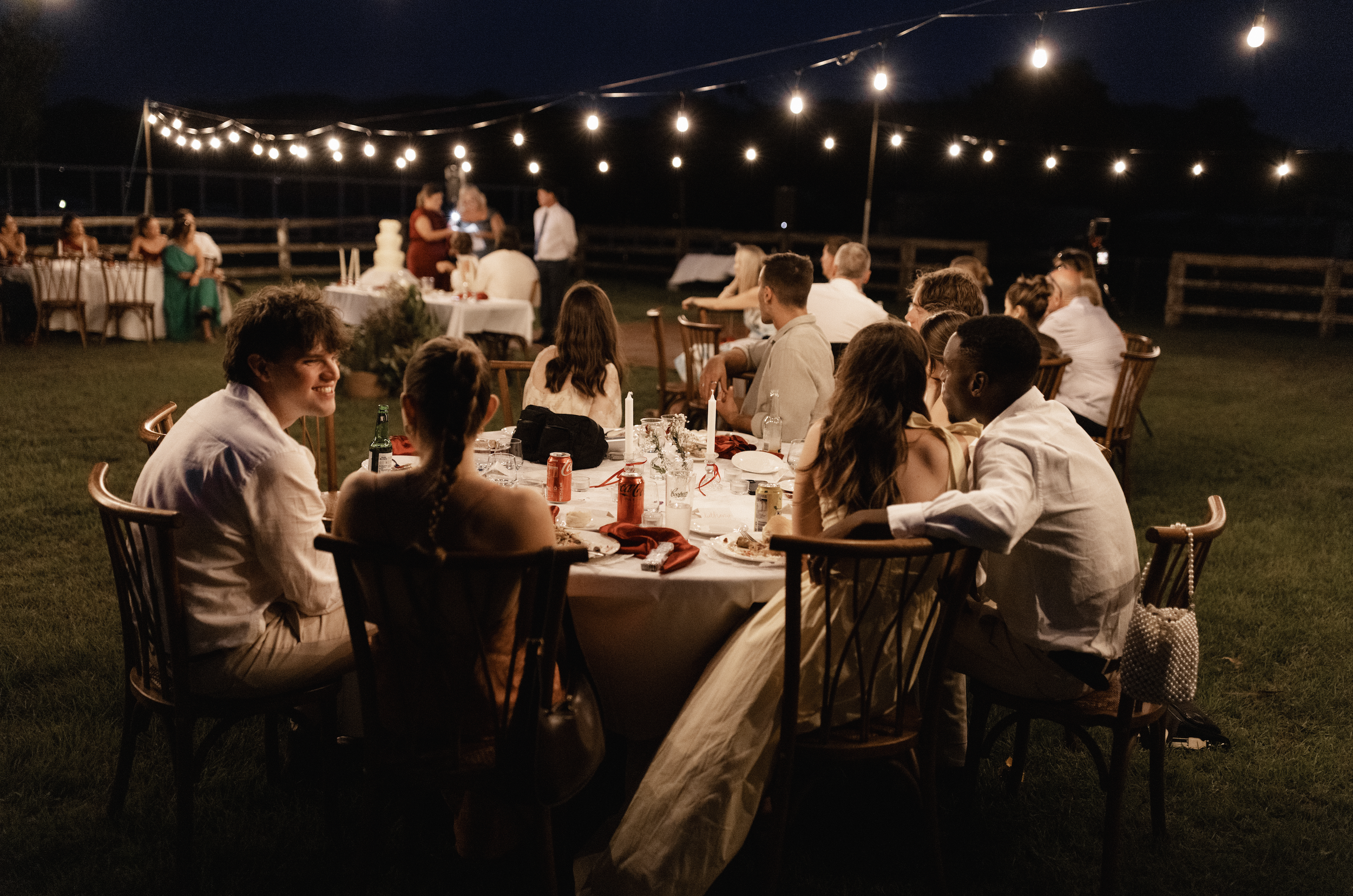 People gathered at a dinner event outdoors at night, sitting around a round table with candles, drinks, and tableware, under string lights.
