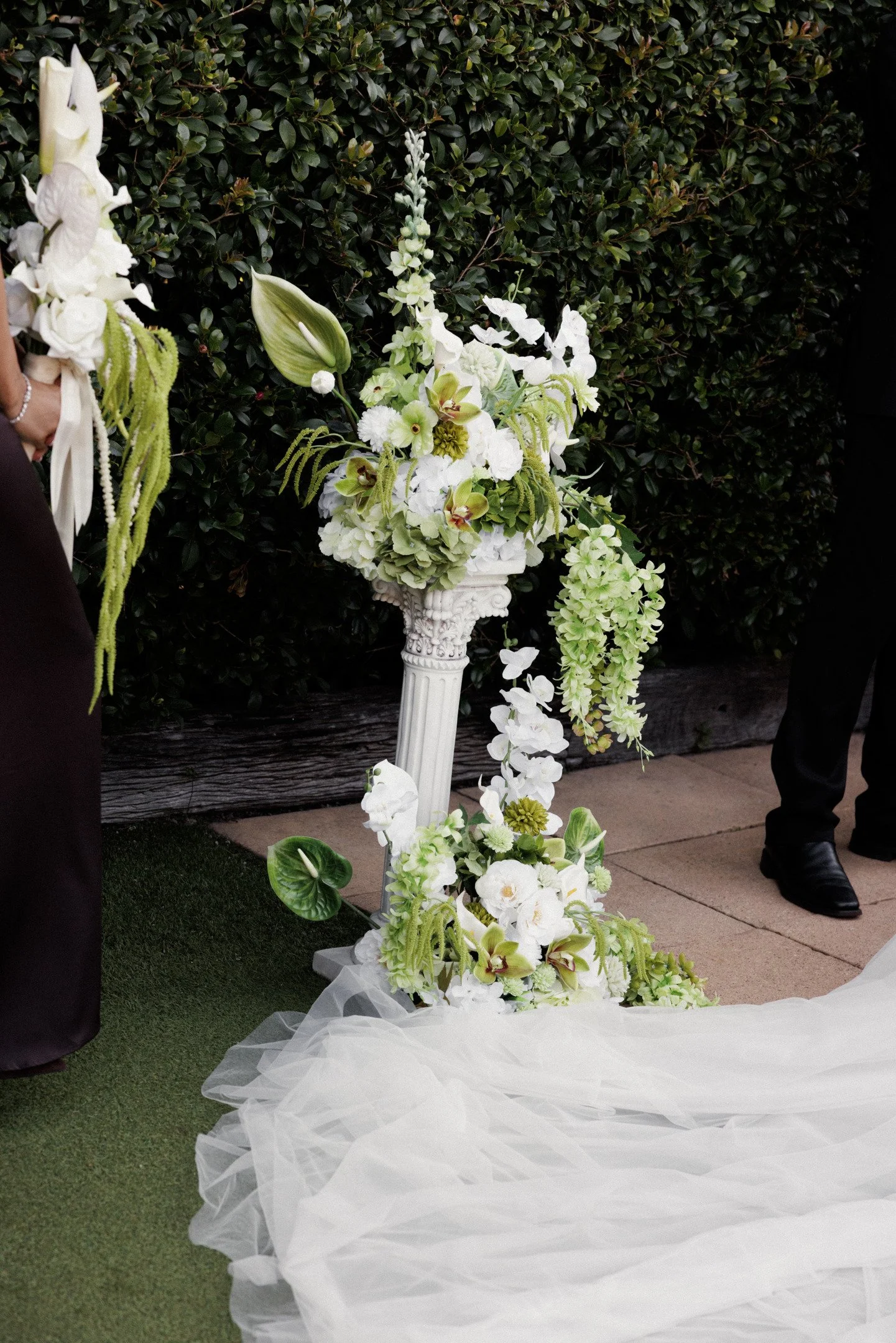 White floral arrangement on a pedestal at a wedding, with white flowers including orchids, roses, and lilies, and draped white fabric on the ground, set against a background of a trimmed shrub.